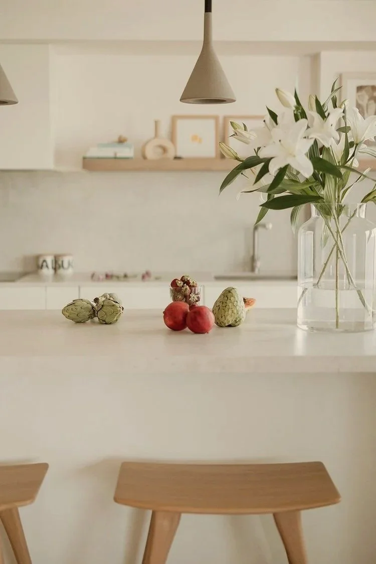 Vaso con flores blancas en la mesa de cocina, con frutas y vegetales como alcachofas, tomate y chayote, fondo con estantes y lámpara.