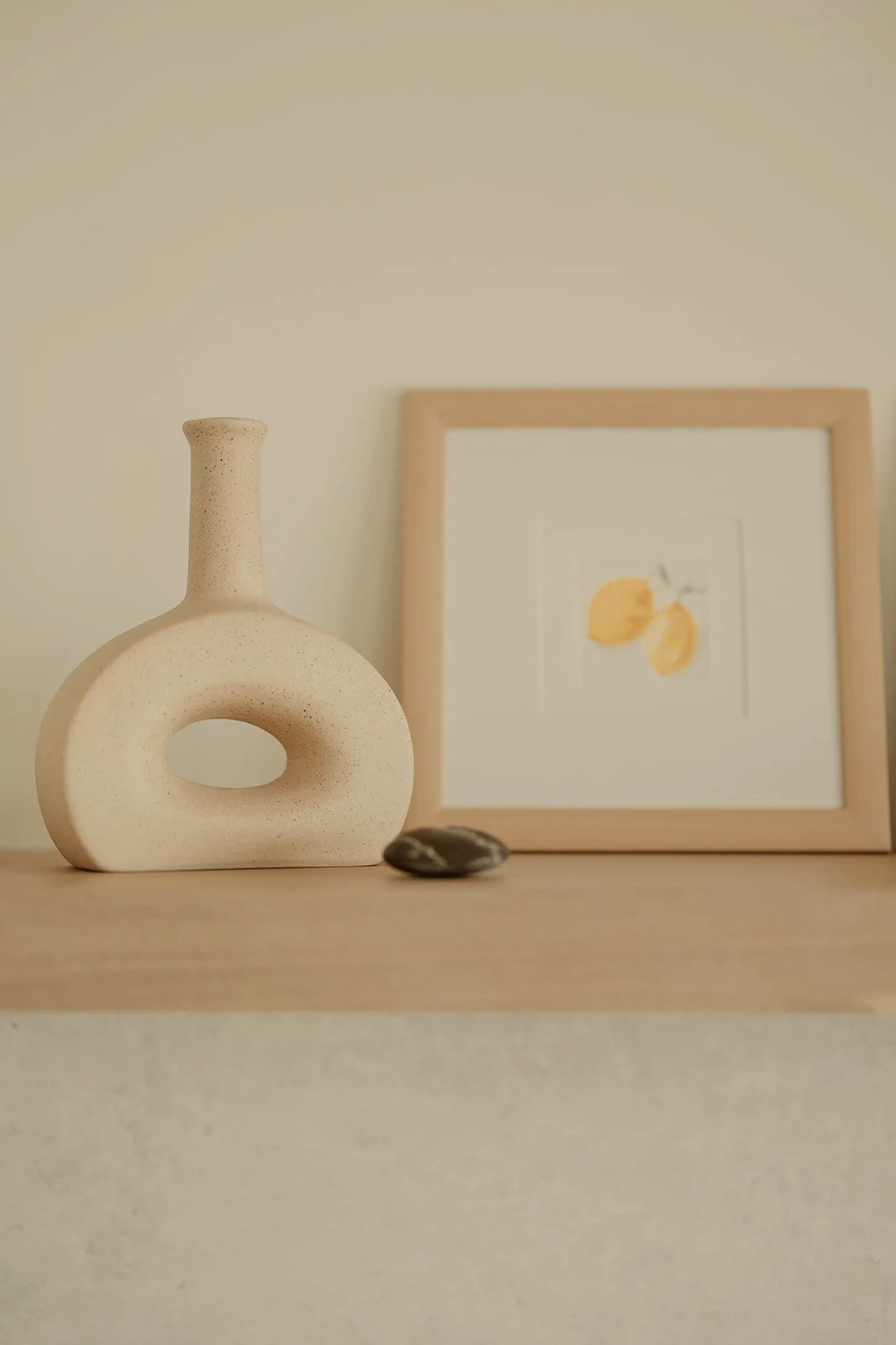 Minimalist beige ceramic decorative vase, framed lemon artwork, and a small black and white stone on a light wood shelf.