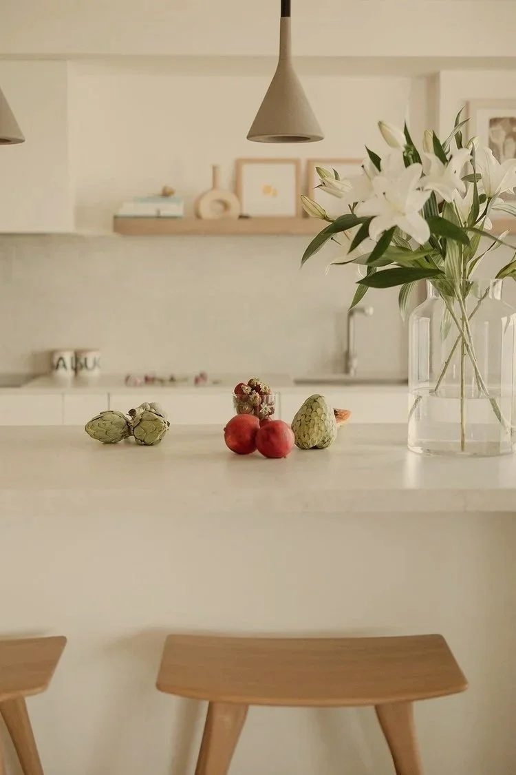 A kitchen countertop with artichokes, peaches, and a bunch of white lilies in a glass vase, with minimalist decor in the background and wooden chairs at the counter.