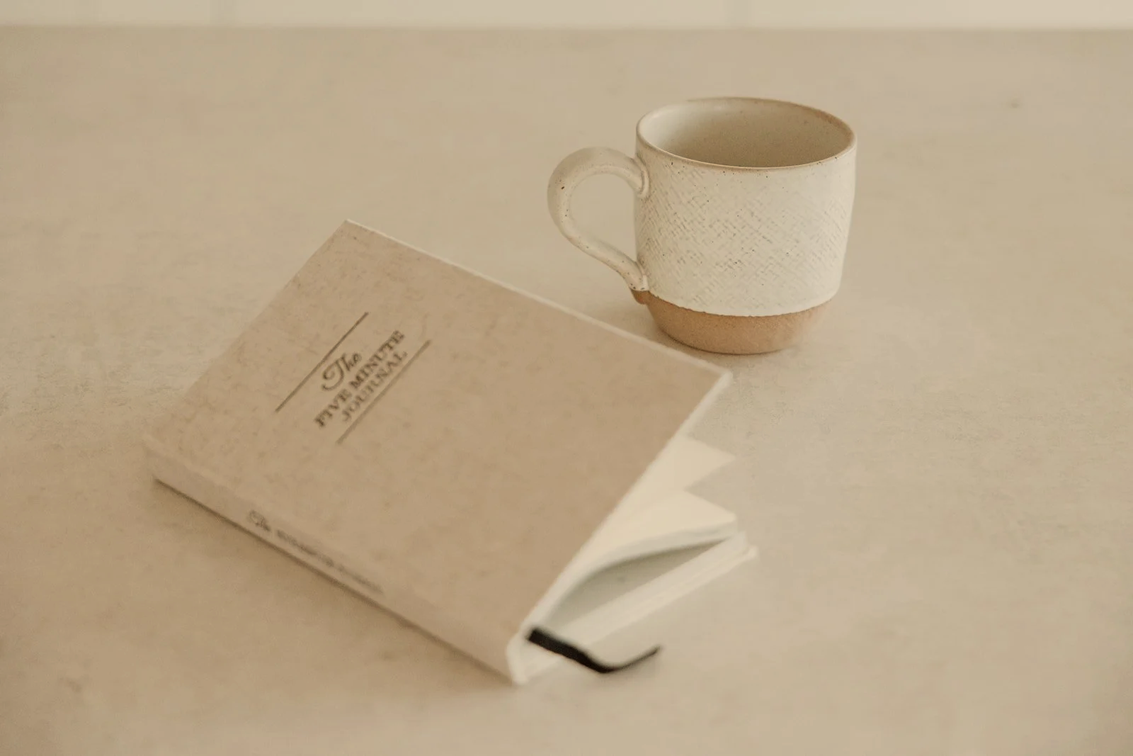 A small white coffee cup and a white book called "The Five Minute Journal" on a plain white surface.