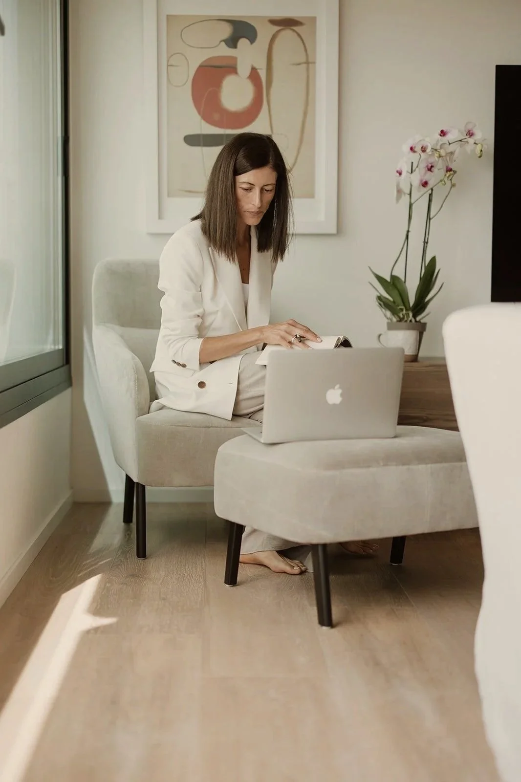 Mujer sentada en una sala de estar, usando una computadora portátil y leyendo un libro, con plantas y arte en el fondo.