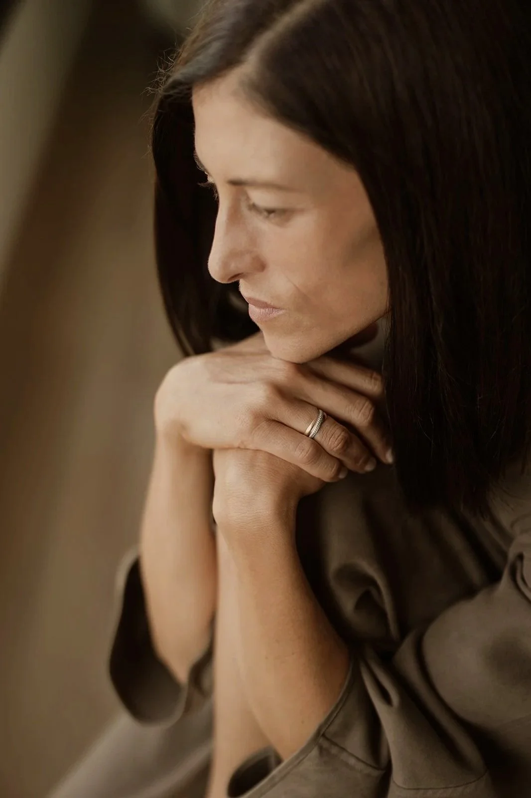 A woman with long dark hair looks down thoughtfully, resting her chin on her hands, wearing a ring on her left hand.