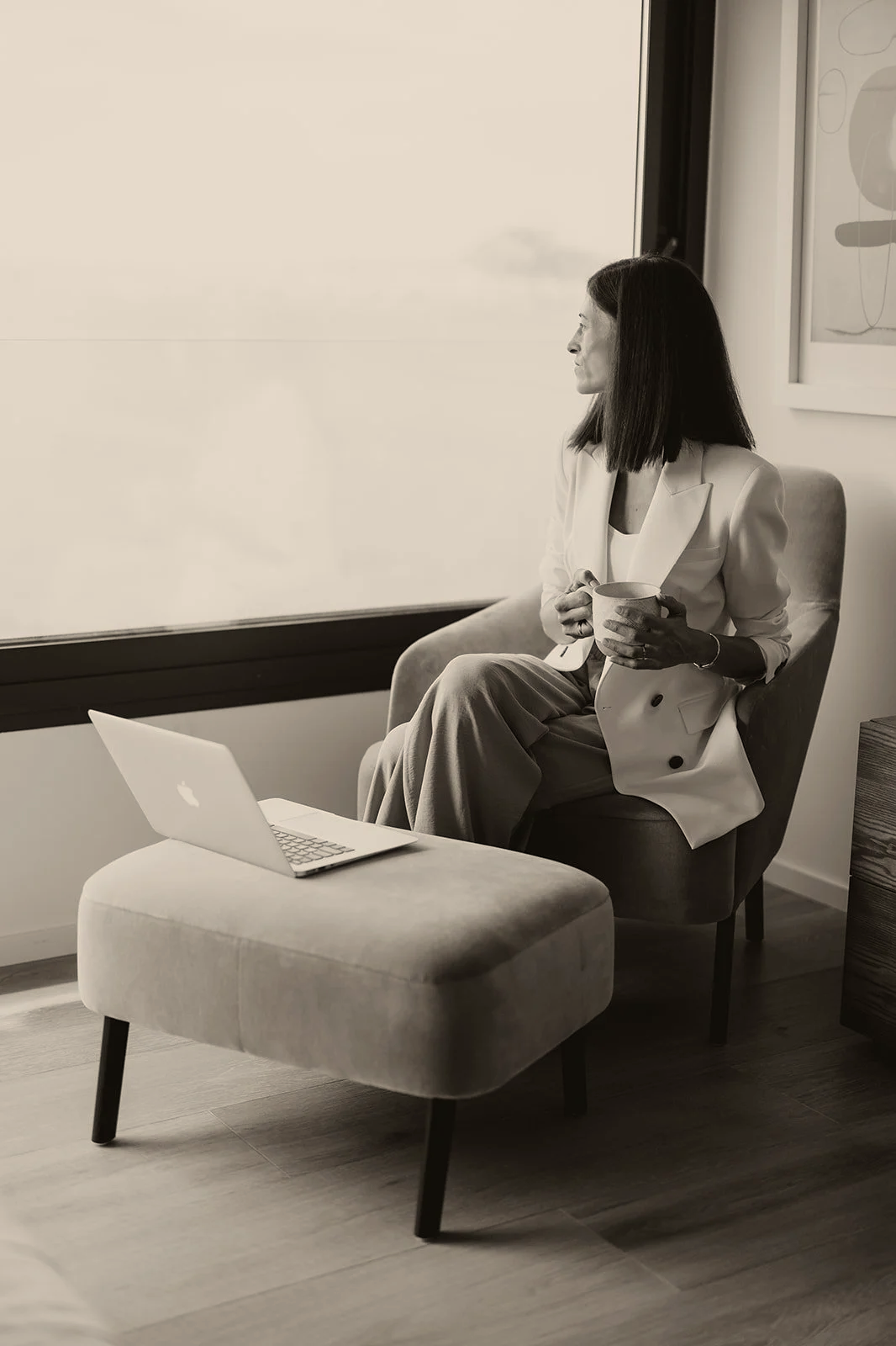 Mujer sentada en una silla, mirando por la ventana, sosteniendo una taza, con una laptop sobre un banco delante de ella.