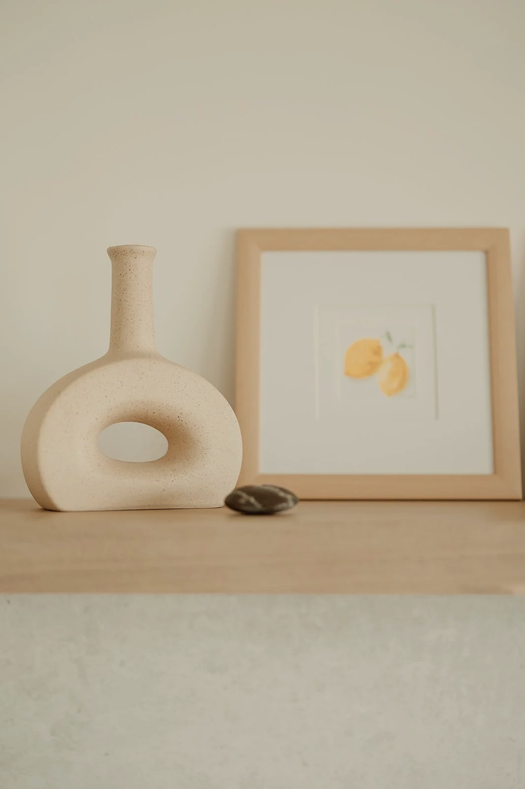Vaso de cerámica beige con forma de figura abstracta, una piedra y un marco con ilustración de limones en el fondo