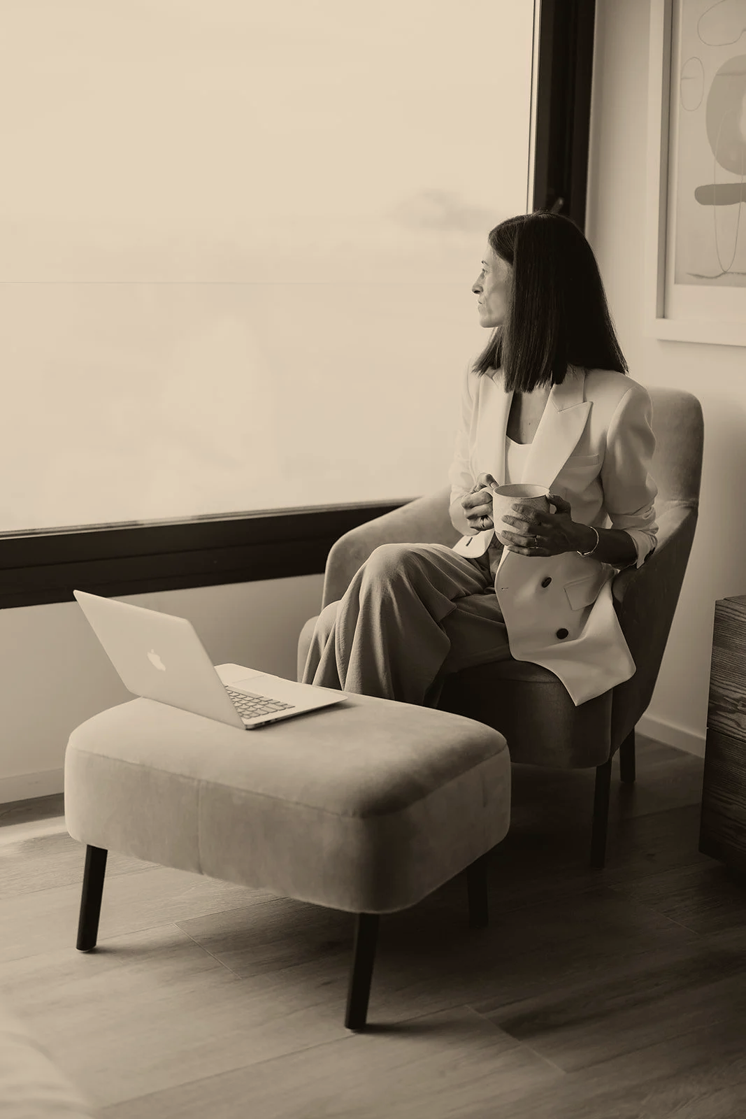 A woman sitting in a chair by a window, holding a mug, with a laptop on an ottoman in front of her, in a cozy indoor setting.
