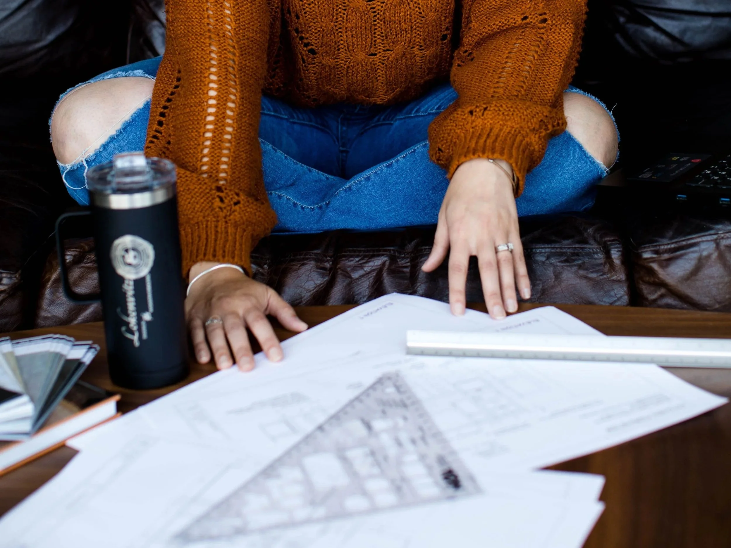 Person wearing a rust-colored knit sweater and ripped jeans sitting cross-legged on a leather couch, working on architectural plans spread out on a wooden table with a ruler and a thermos nearby.