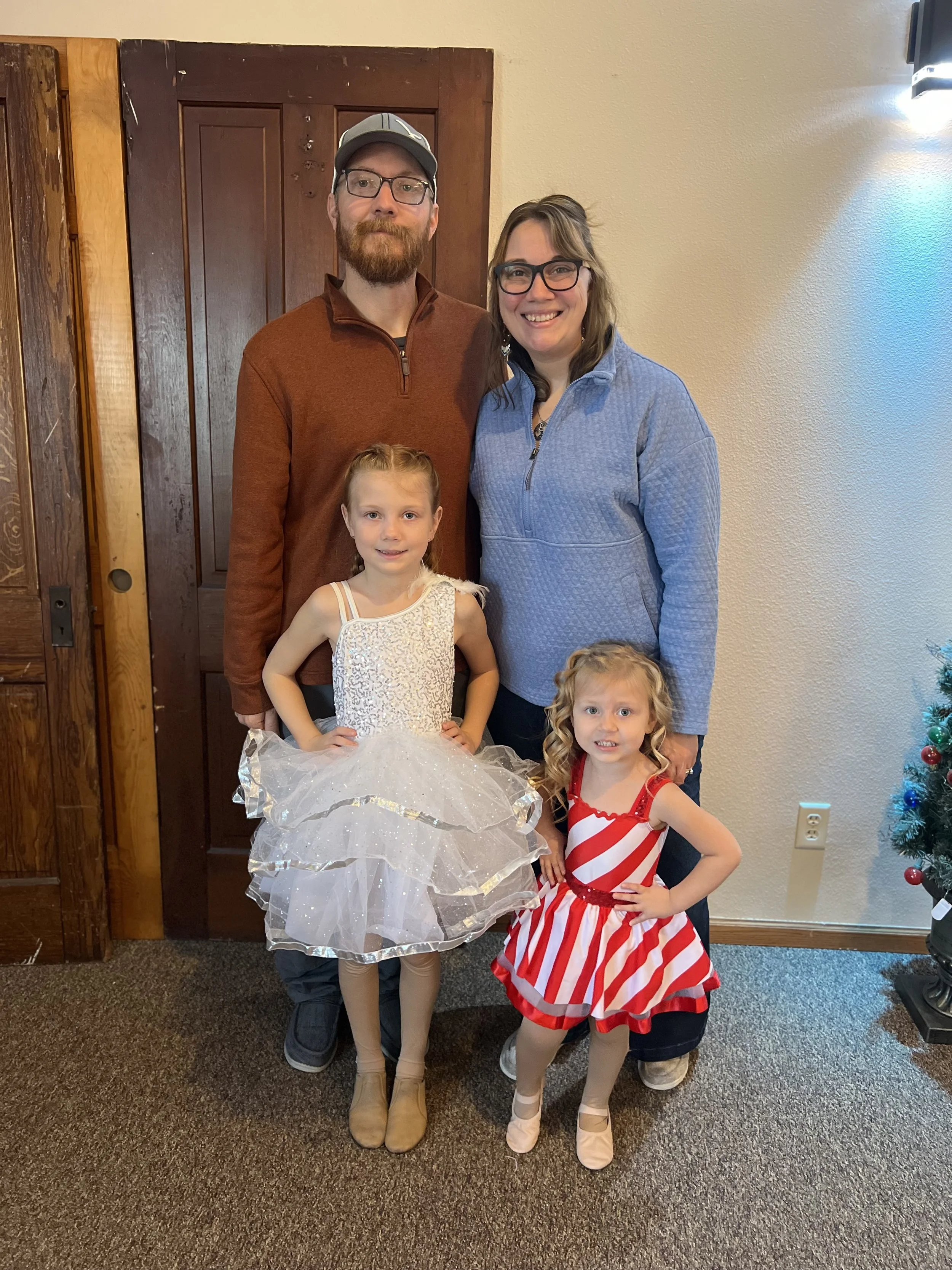A family of four posing indoors in front of a wooden door and wall, with a decorated Christmas tree on the right. The father has a beard, glasses, a gray cap, and wears a brown zip-up jacket. The mother has glasses, shoulder-length brown hair, and wears a blue zip-up sweater. The two young girls are wearing dresses: the older girl in a white, sparkly dress with a tulle skirt, and the younger girl in a red and white striped dress. All are smiling and standing close together.