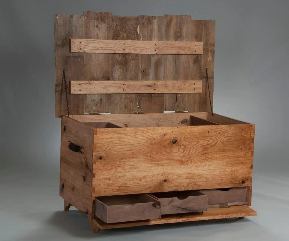 A wooden chest with a lid that is open, revealing two compartments inside. The chest has a rustic, unfinished look with visible wood grain, knots, and metal hinges. There are two small drawers at the bottom front of the chest.