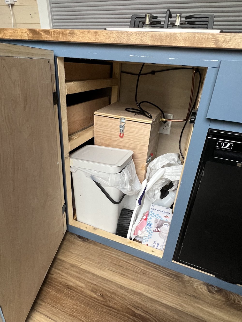  Cabinet in van camper showing propane box, garbage can, and other cleaning items. 