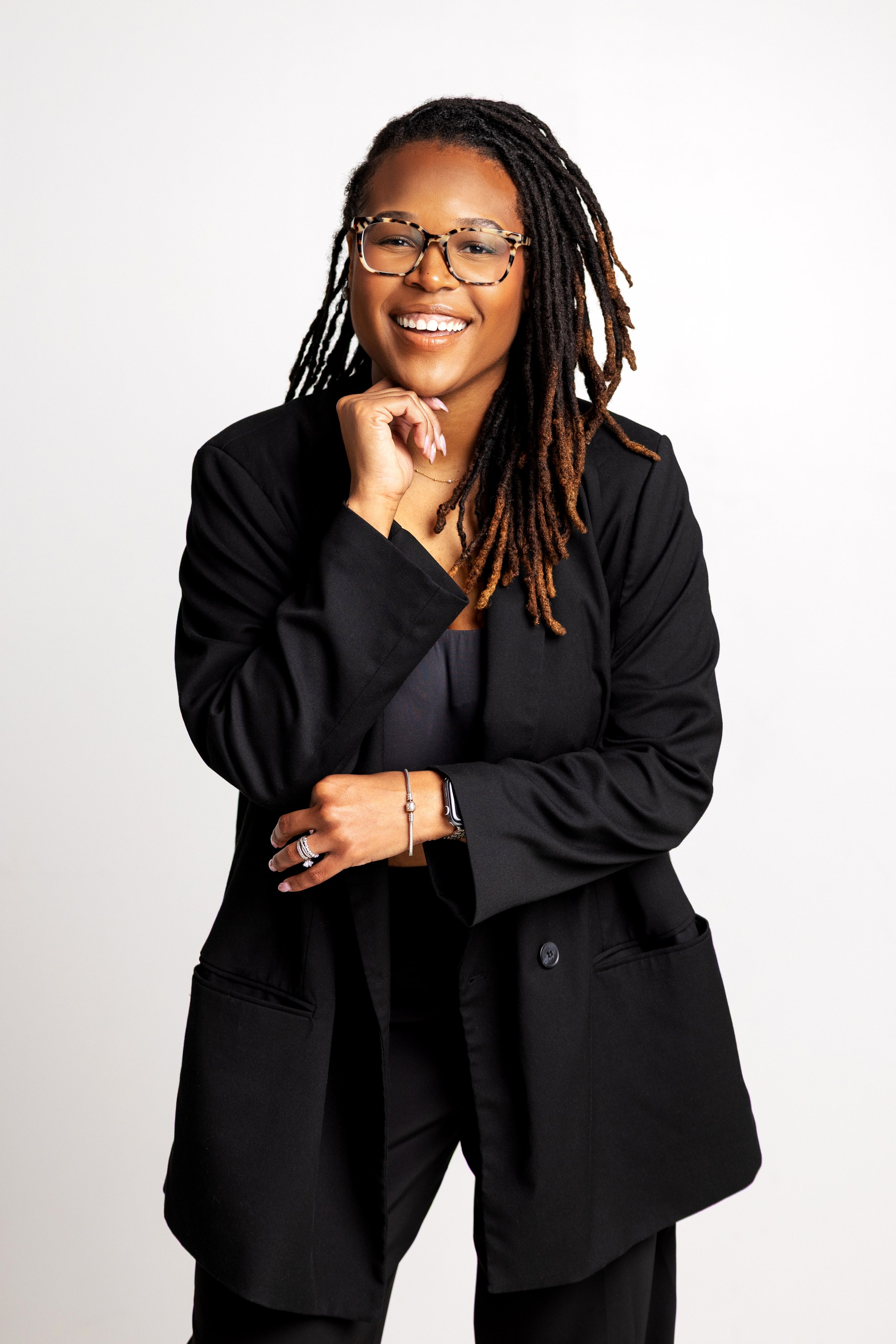 A confident woman with dreadlocks and glasses smiling, wearing a black blazer and statement jewelry, standing against a plain white background.