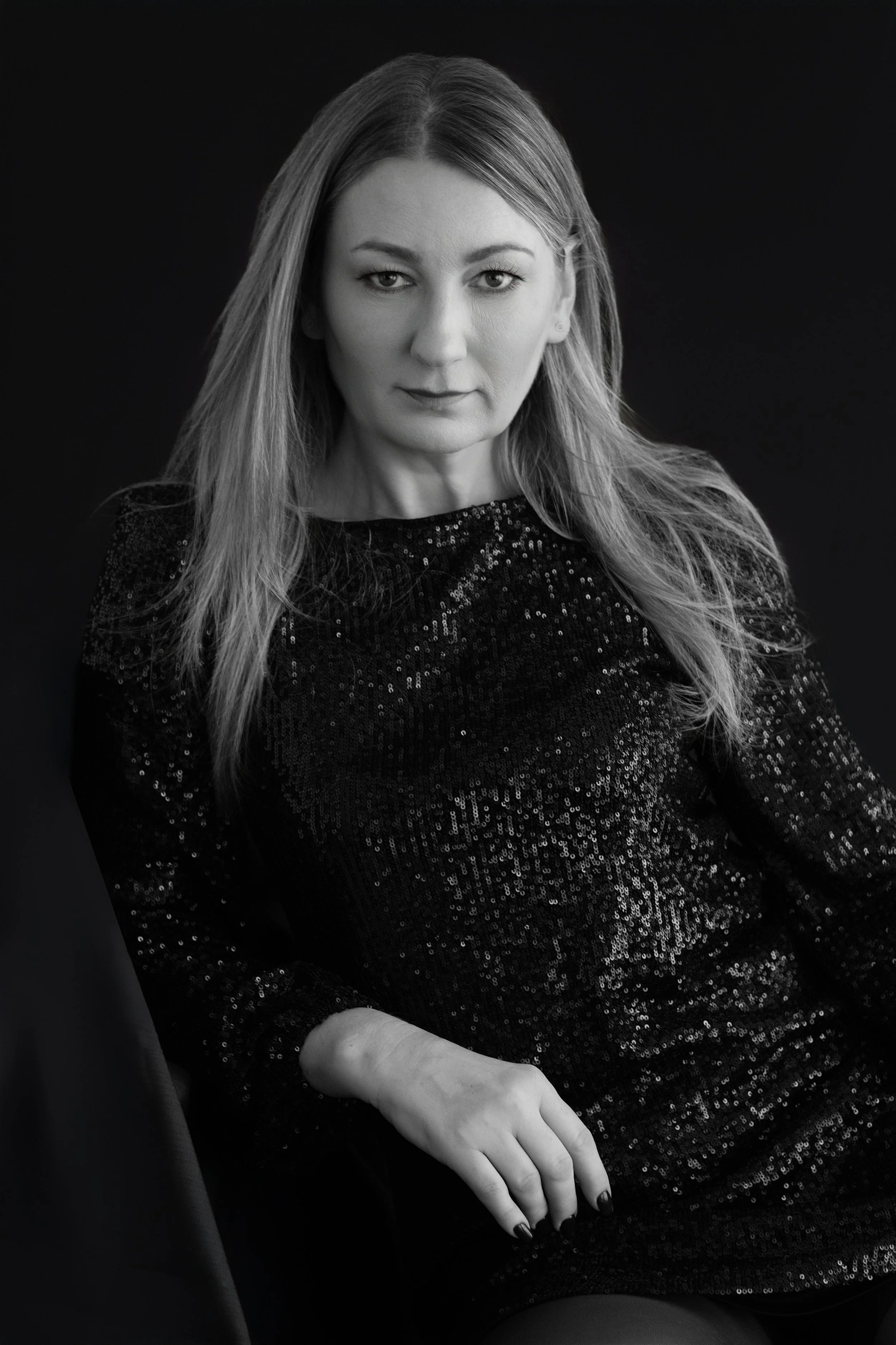 Black and white portrait of a woman with long hair, wearing a sequined dress, seated against a dark background.