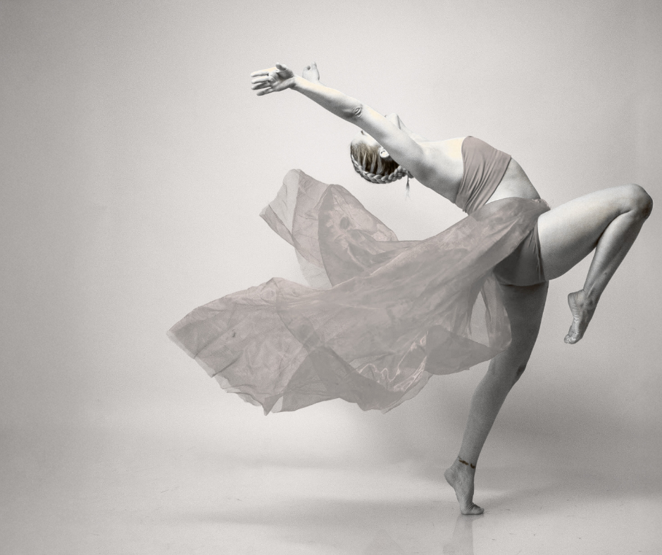 A woman dancing or performing a pose, wearing a flowing skirt and sports bra, with her arms extended and one leg raised, standing on one foot against a plain background.