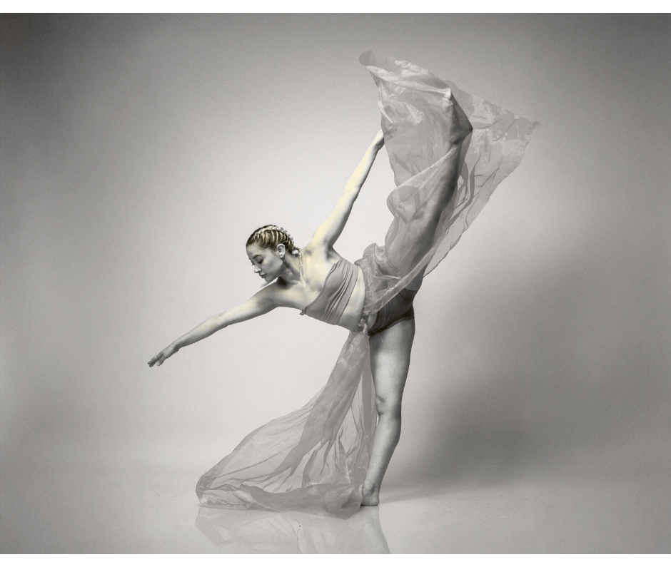 A woman ballet dancer performing a pose with one leg lifted high and one arm extended, wearing a flowing skirt, in a dance studio or photo studio with a plain background.