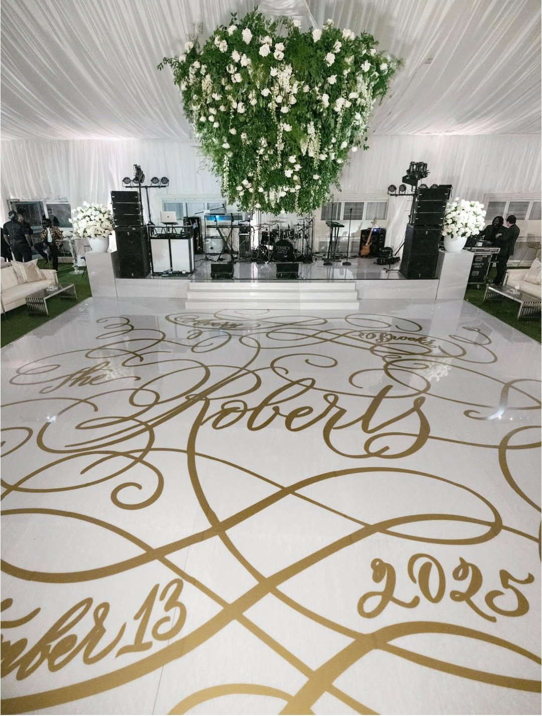 Elegant wedding venue with a stage, floral arrangements, and a white dance floor with gold calligraphy reading 'The Roberts' and details of the wedding date, October 13, 2025.