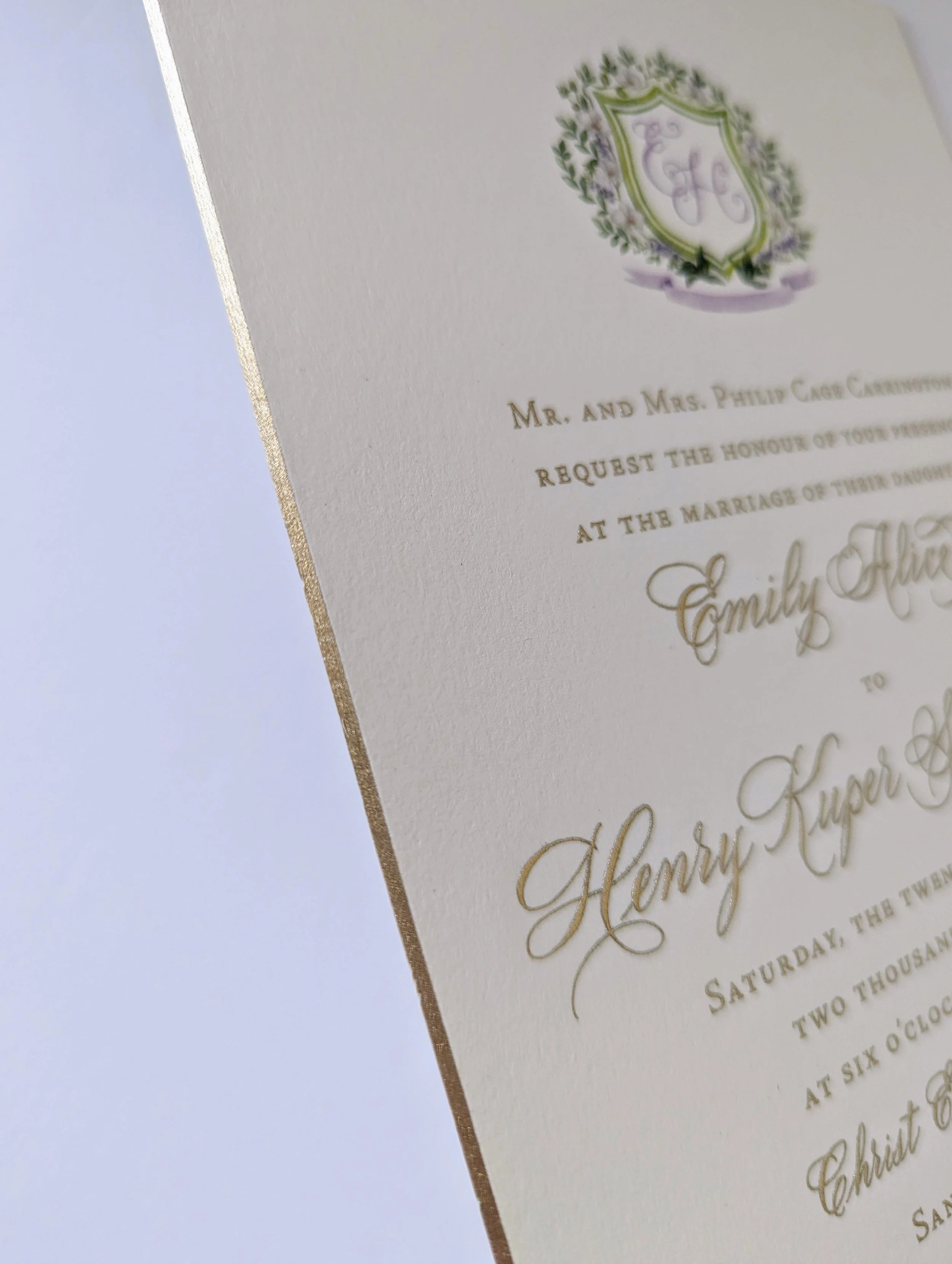 Close-up of a wedding invitation card with gold foil details, featuring a decorative crest with a shield and floral design, text in elegant script and serif fonts, partially visible event details, and a gold border on the left edge.