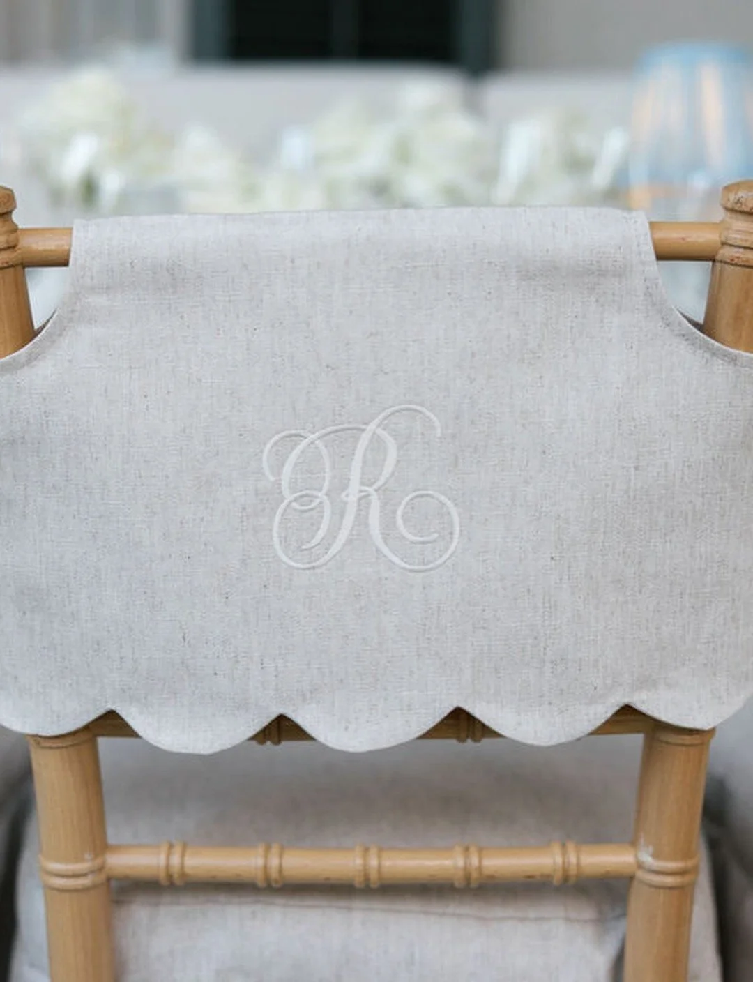 A white fabric cover with embroidered initials 'R' on a wooden chair with a scalloped edge, set in a blurred background of a dining area.