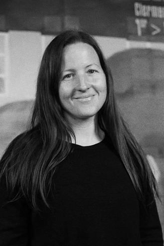 A woman with long dark hair smiling at the camera, wearing a black top, indoors