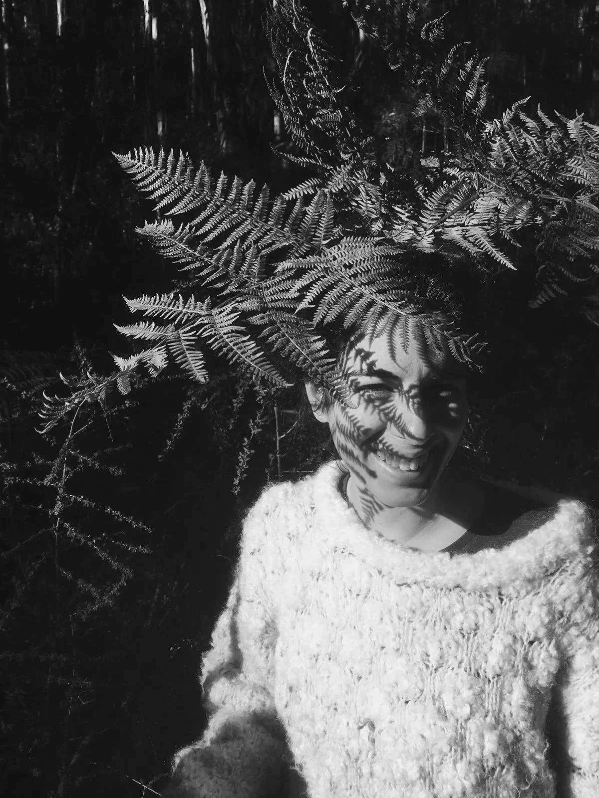 A smiling person in a textured sweater, mostly covered in shadow with fern leaves casting shadows on their face, in a dark outdoor setting.