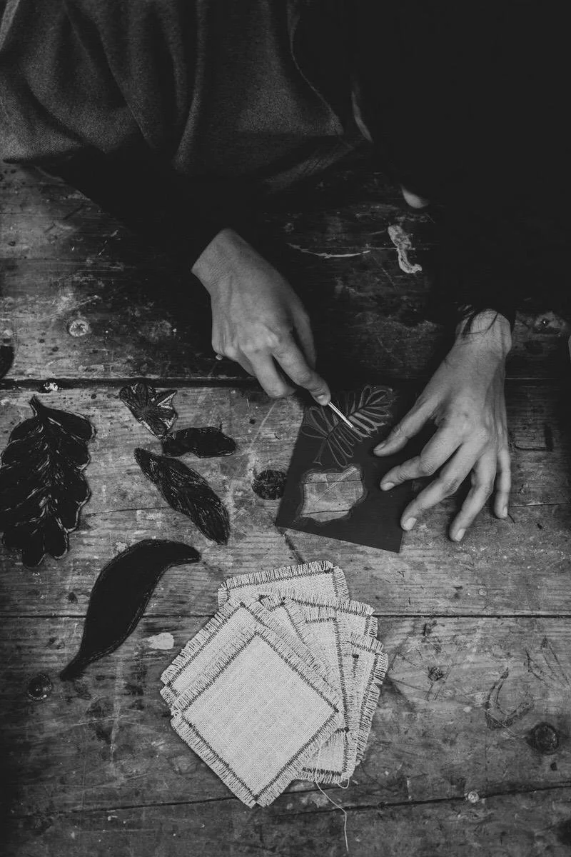 Person creating rub-on adhesive leaves and plants design on a piece of paper or fabric, with various leaves and fabric pieces on a wooden table.