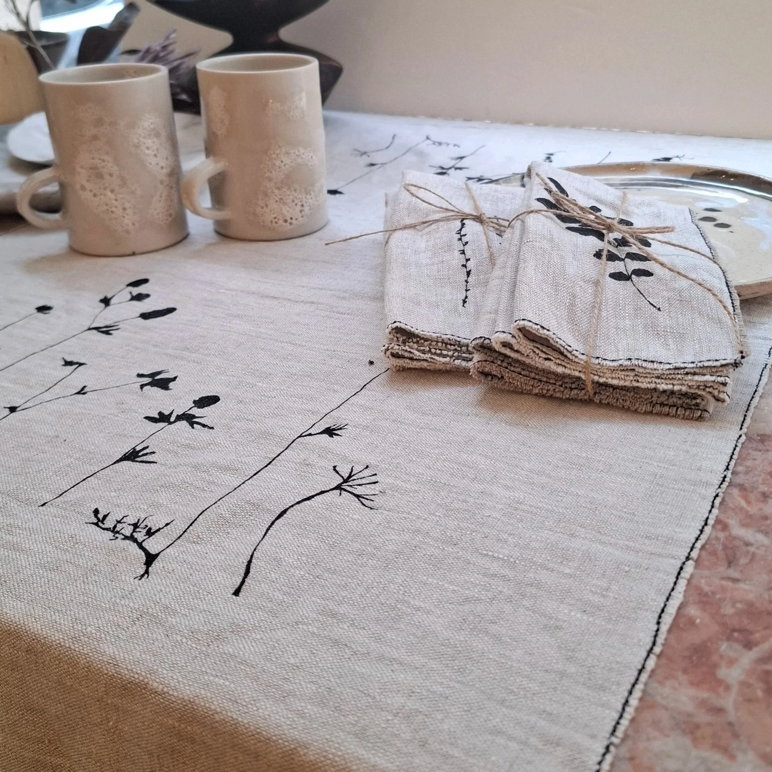 A table setting with beige mugs, folded linen napkins tied with twine, and a serving tray on a fabric tablecloth with black botanical prints.