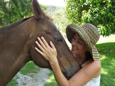 A woman smiling while petting a brown horse outdoors in a lush green setting.
