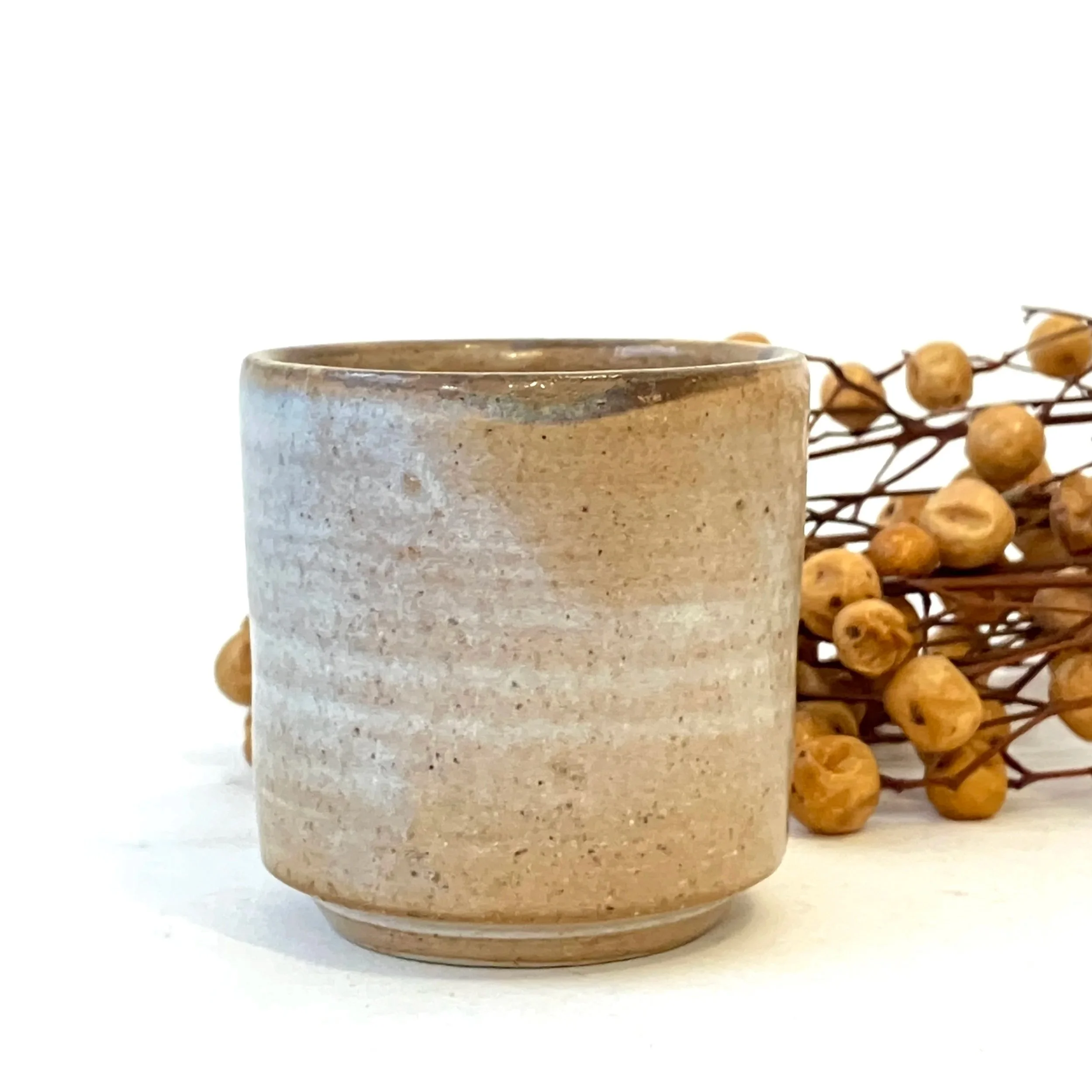 Beige ceramic mug with a slightly textured surface, positioned beside a branch of dried yellow berries on a white background.