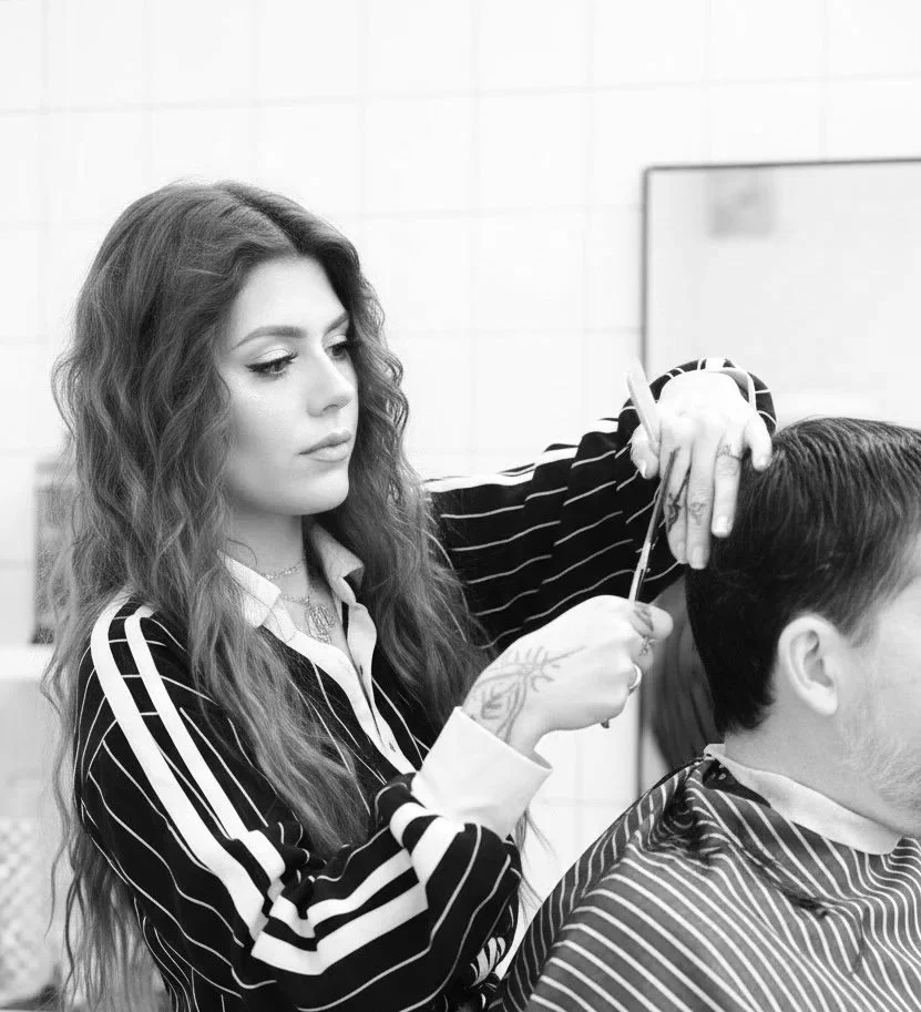 A woman with curly hair and braids is smiling while styling a man's short hair in a salon.