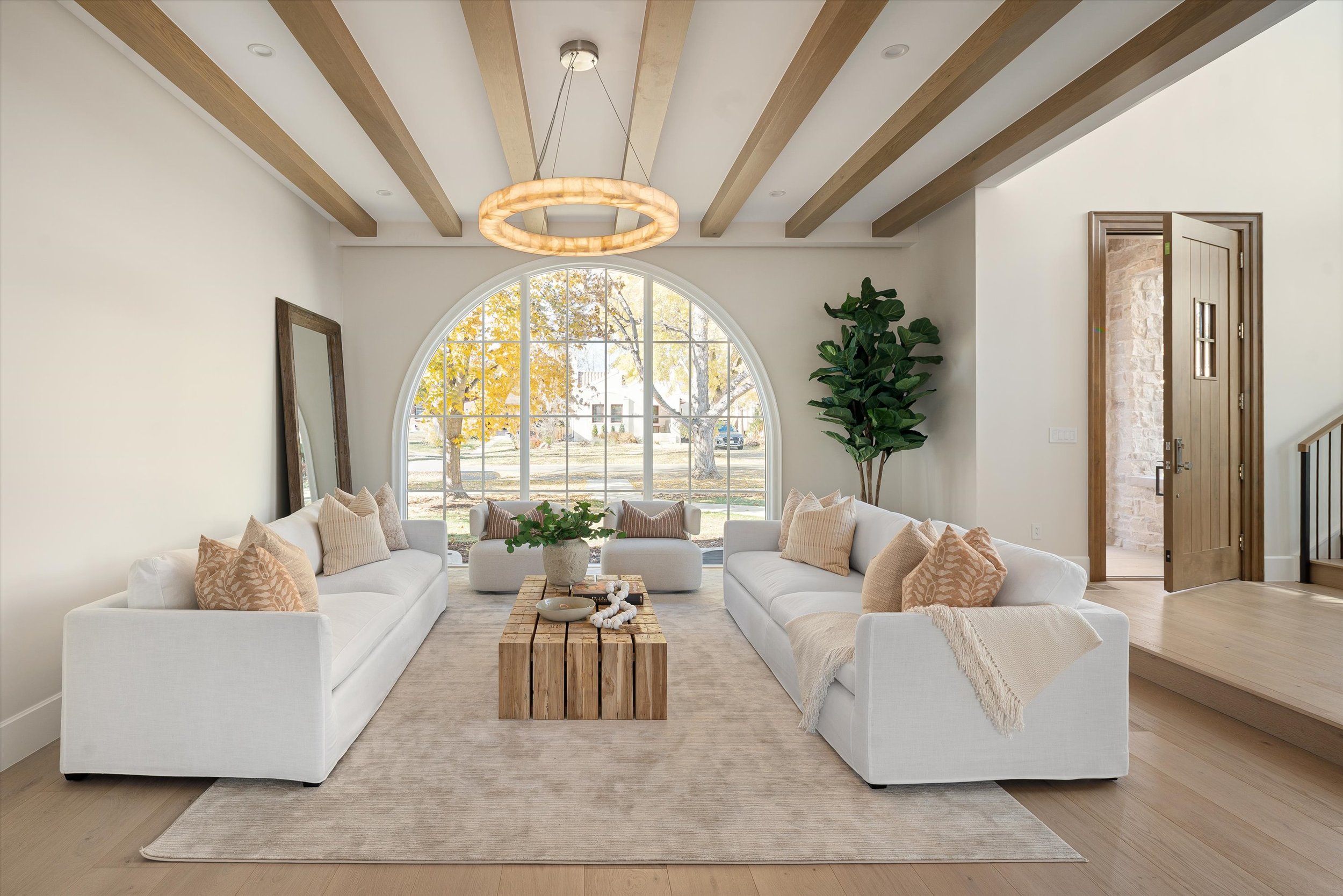 Living room with two white sofas, wooden coffee table, large arched window with trees outside, beige rug, interior plants, and wooden accents.