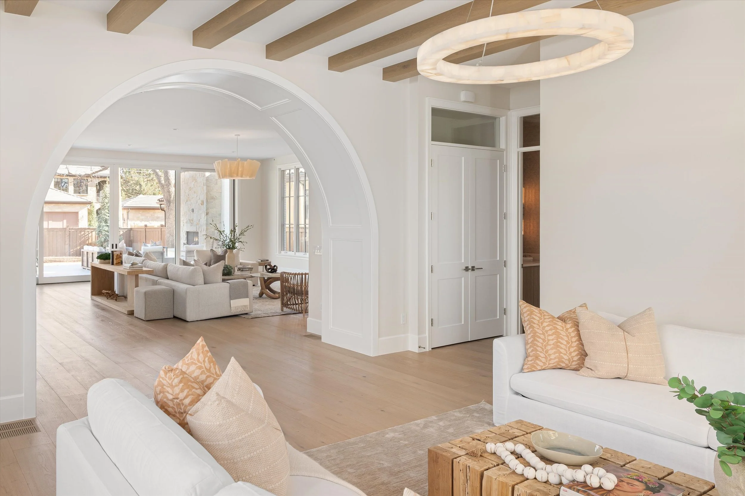 Bright living room with white sofas, beige and peach pillows, wooden coffee table with decorative items, and large windows with a view of a backyard.
