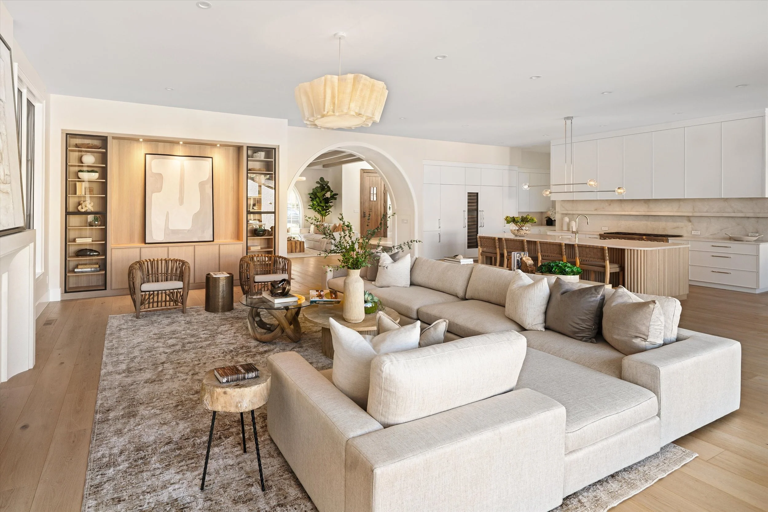 Open-concept living room with beige sectional sofa, wooden chairs, a glass coffee table, and built-in shelves with art and decorative objects. The space flows into a white kitchen with an island and bar stools, and a doorway with a view of a cozy sit