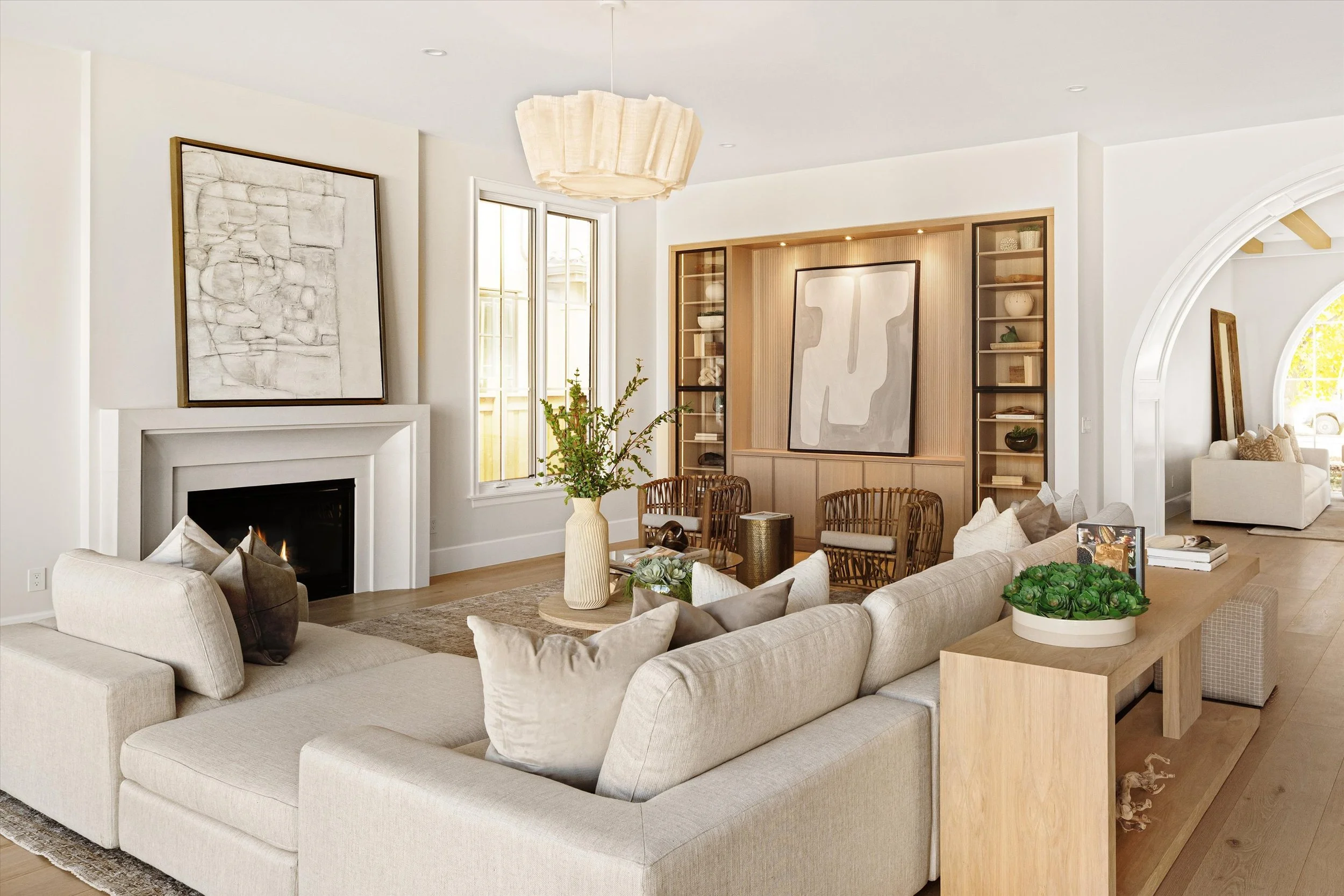 Bright living room with white walls, beige sofa, fireplace, large window, wooden accents, wicker chairs, decorative plants, abstract artwork, and modern light fixture.
