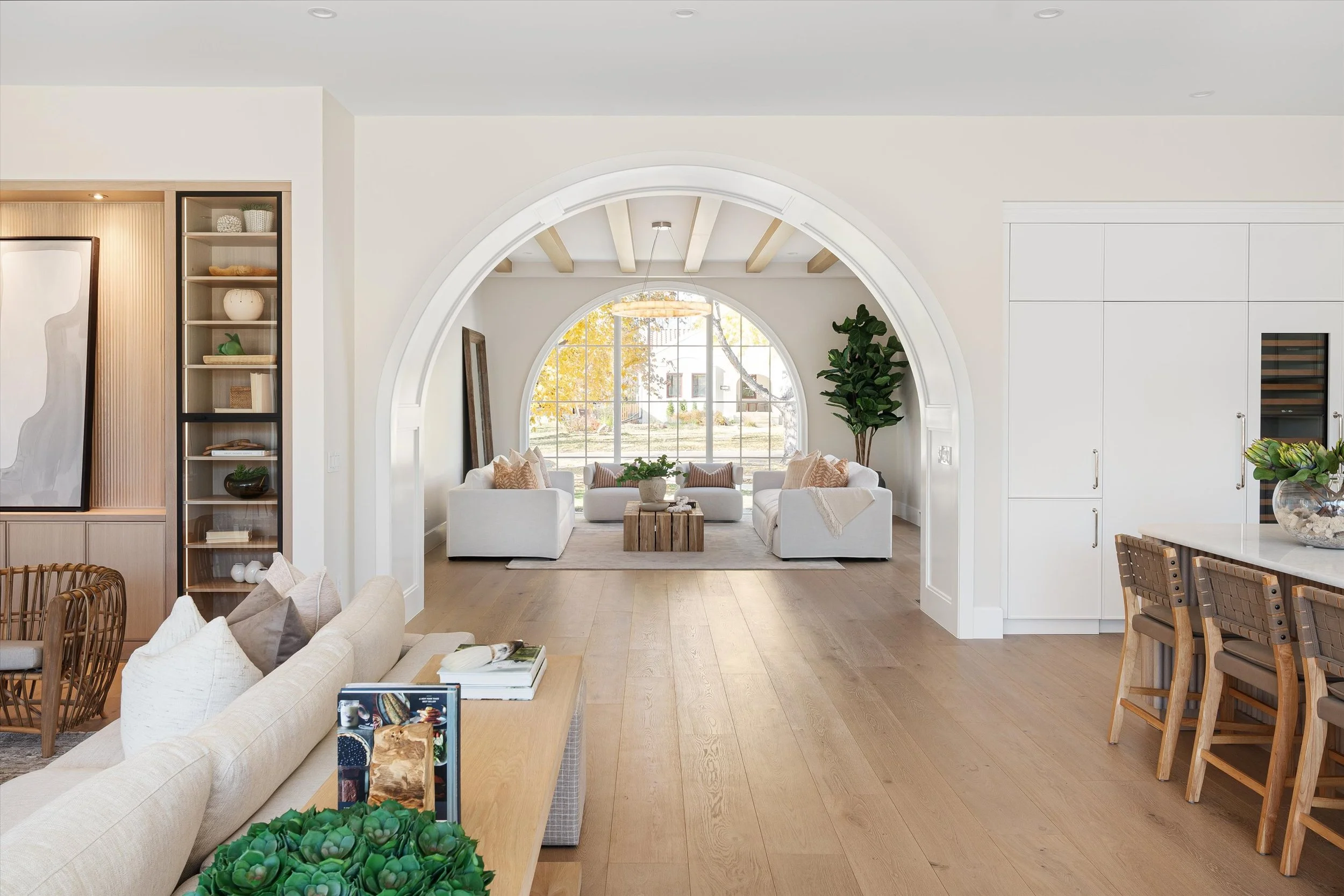 Bright, modern living room with white sofas, large arched window, wooden flooring, and various decorative items and plants.