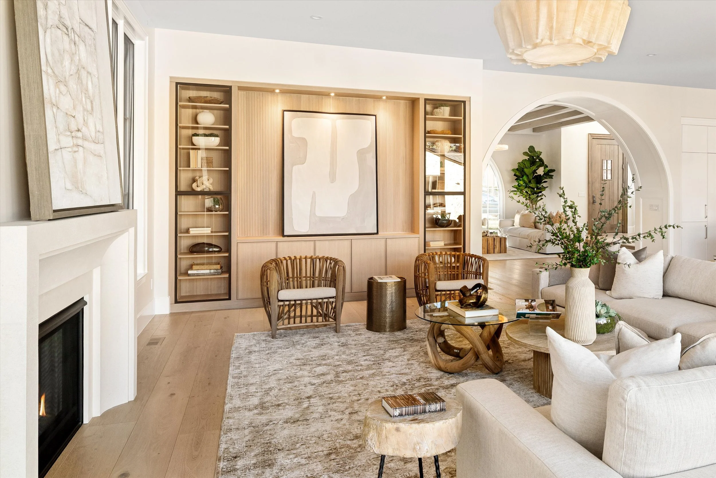 Living room with built-in light wood shelves, artwork, beige couches, rattan chairs, a glass coffee table, a large potted plant, a fireplace, and an arched doorway.