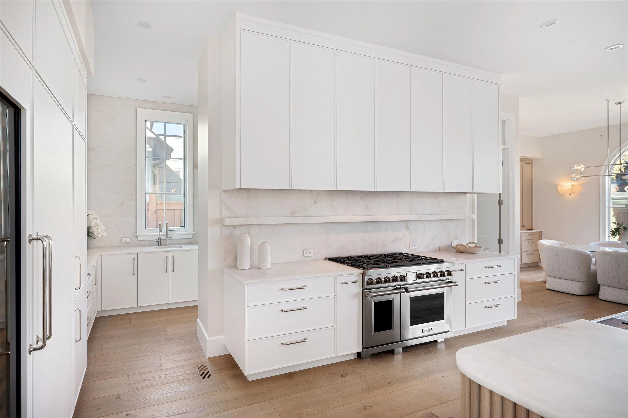 A modern kitchen with white cabinets, a marble backsplash, and a stainless steel oven. Light wood flooring and a window above the sink are visible.
