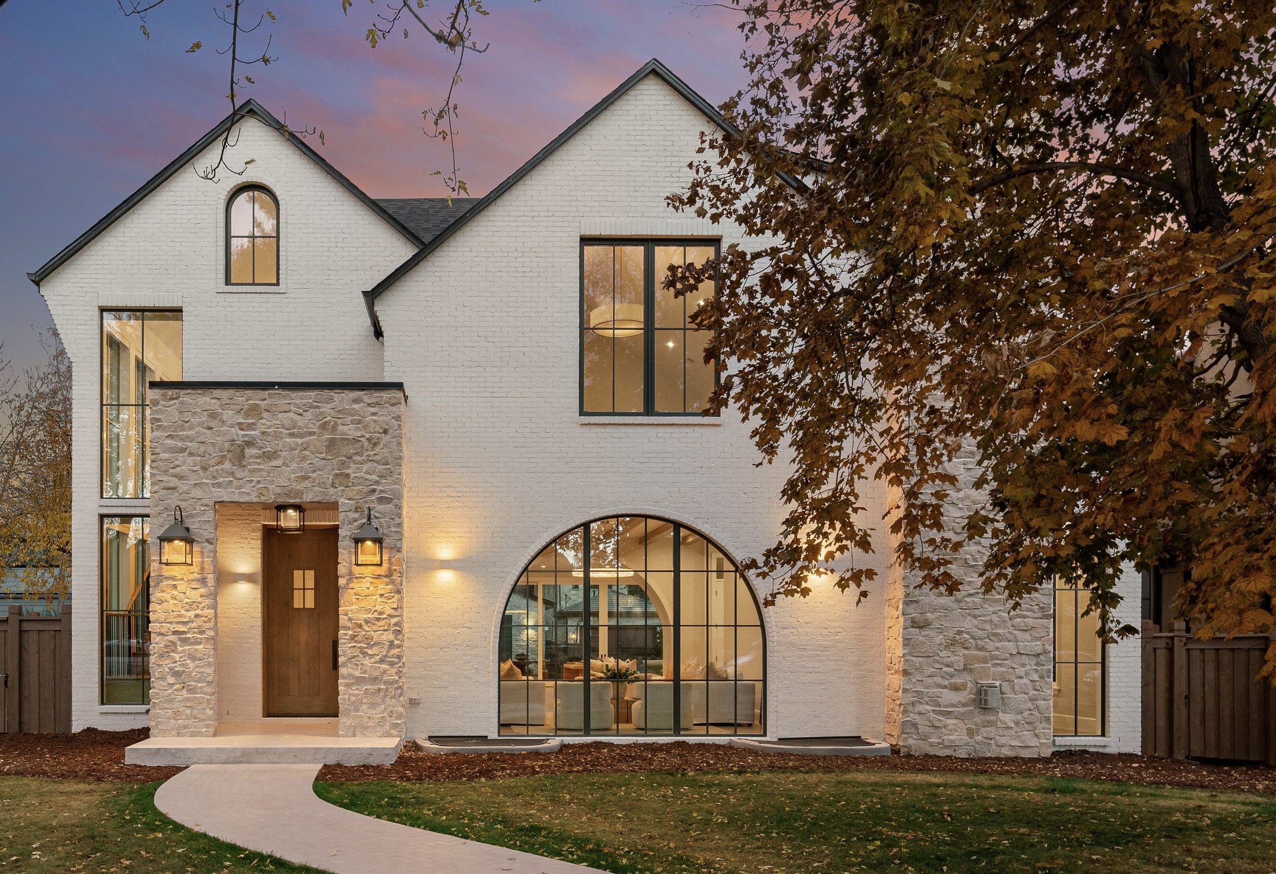 A modern two-story house with white brick walls, a stone accent on the entrance, large black-framed windows, and warm exterior lighting, surrounded by trees with autumn leaves.