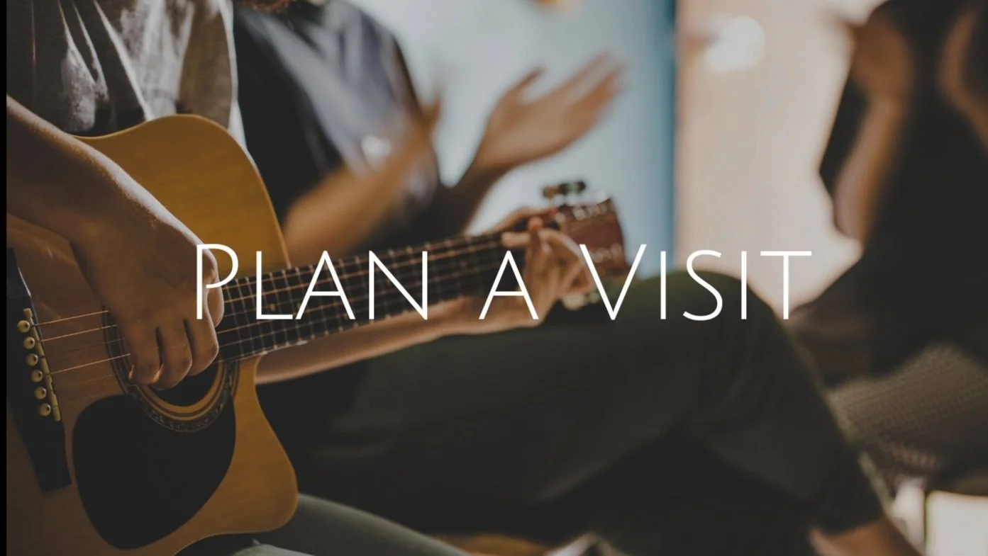 A person playing an acoustic guitar during a gathering with others, with the text "Plan A Visit" overlayed.