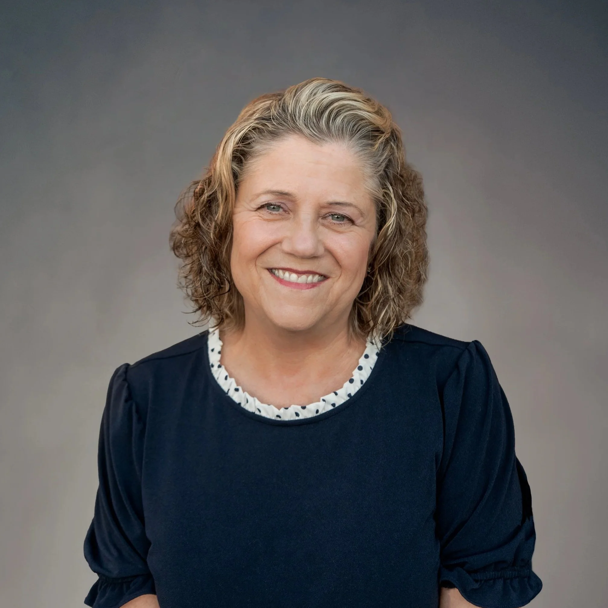 A professional woman with shoulder-length curly hair, smiling, wearing a black and white patterned blazer, pearl earrings, and a necklace, against a neutral gradient background.