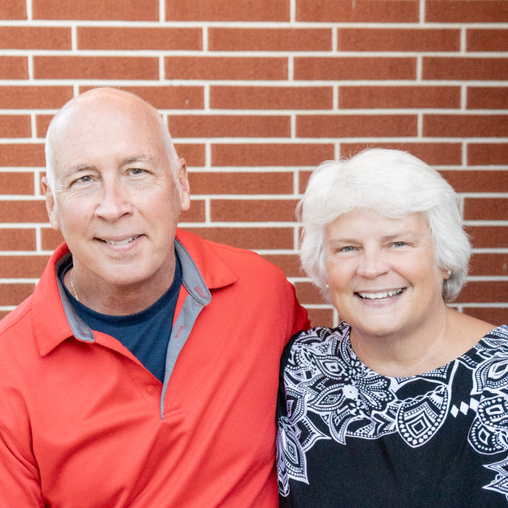 A smiling middle-aged man and woman standing in front of a brick wall, close together.