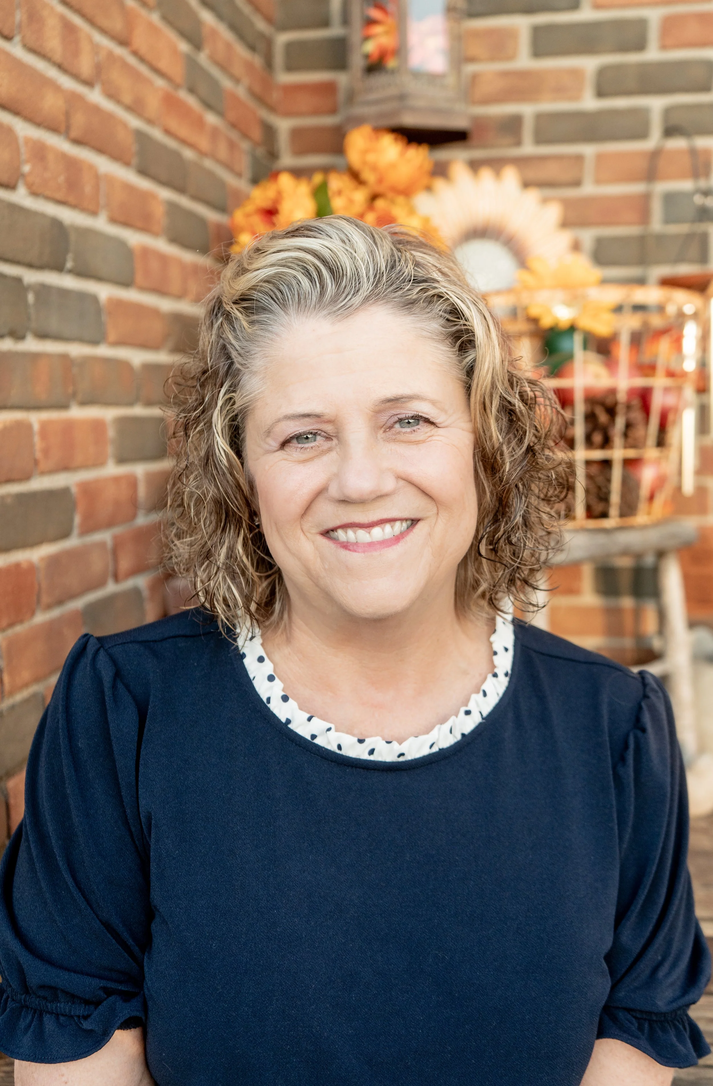A smiling middle-aged woman with curly blonde hair sitting against a brick wall with fall-themed decorations in the background.