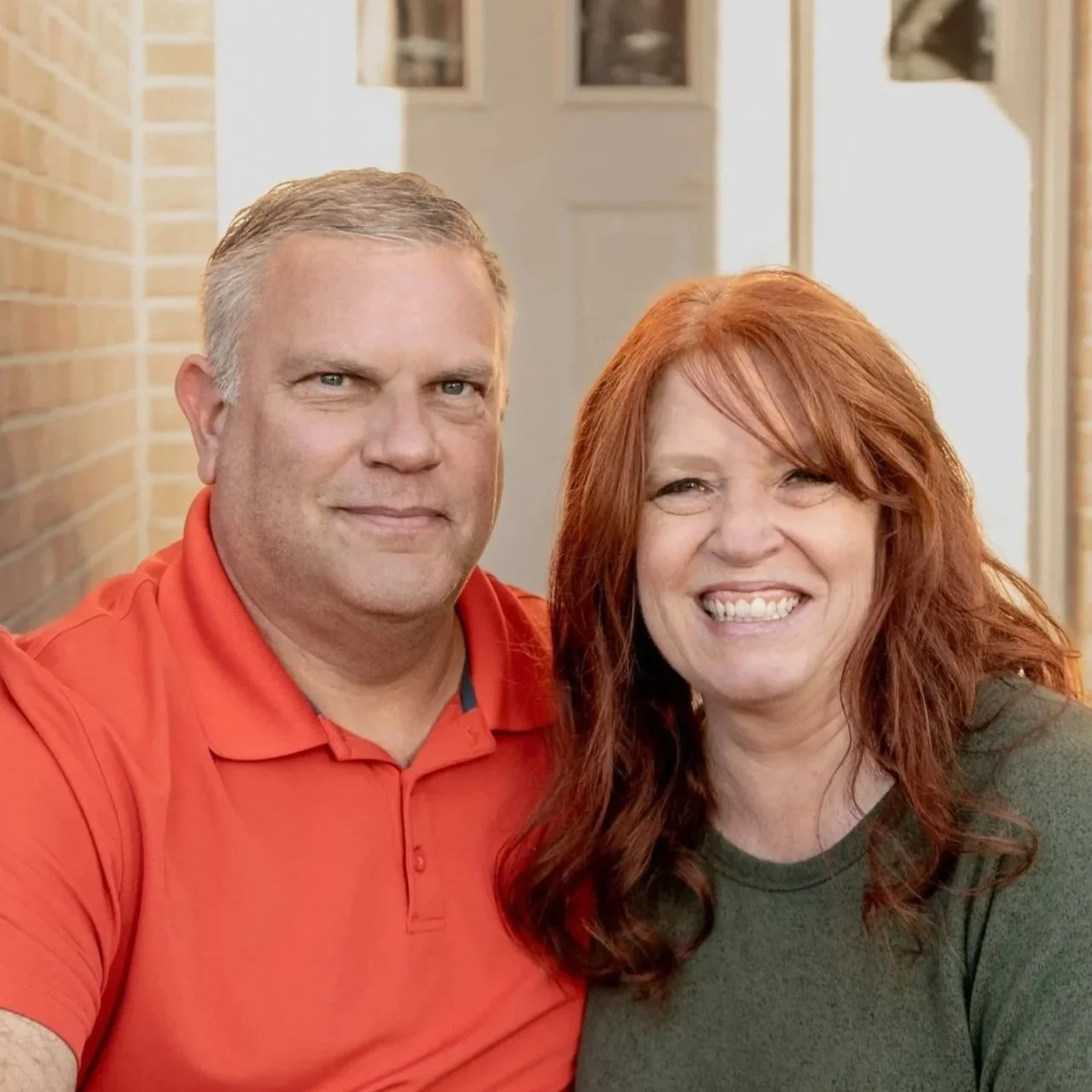 A smiling man and woman sitting outdoors in front of a house, brick wall, and doorway.