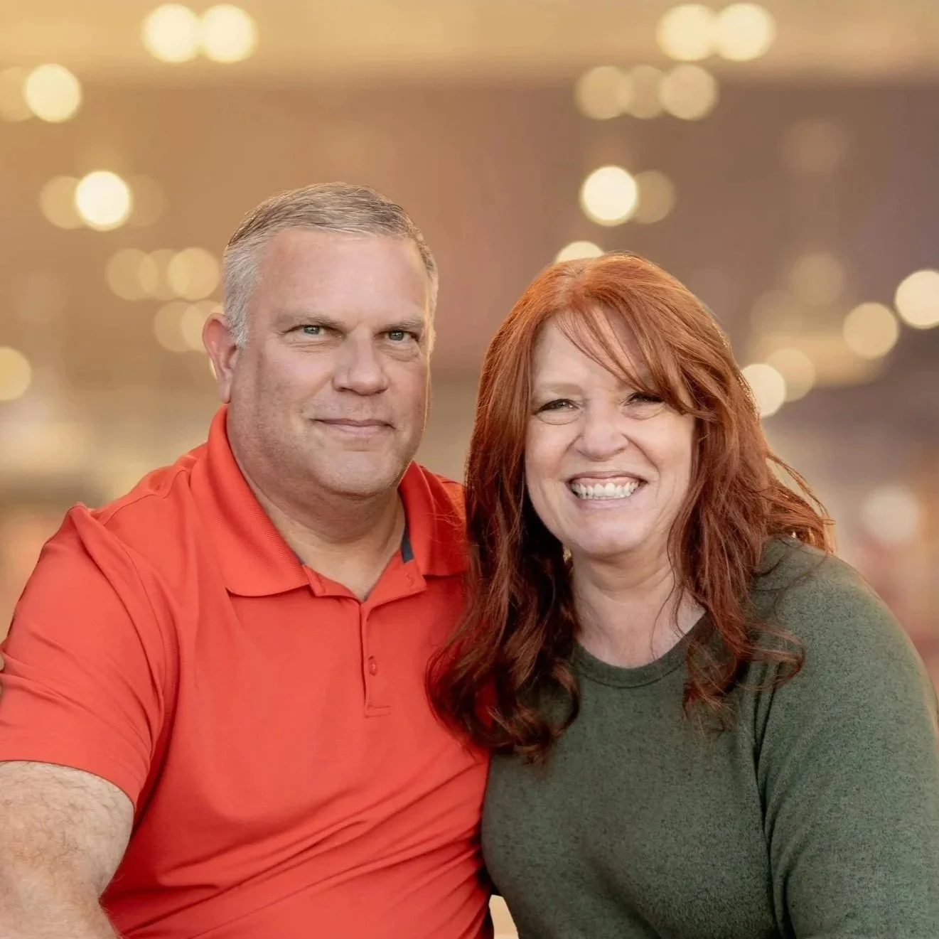 A smiling man and woman sitting outdoors in front of a house, brick wall, and doorway.