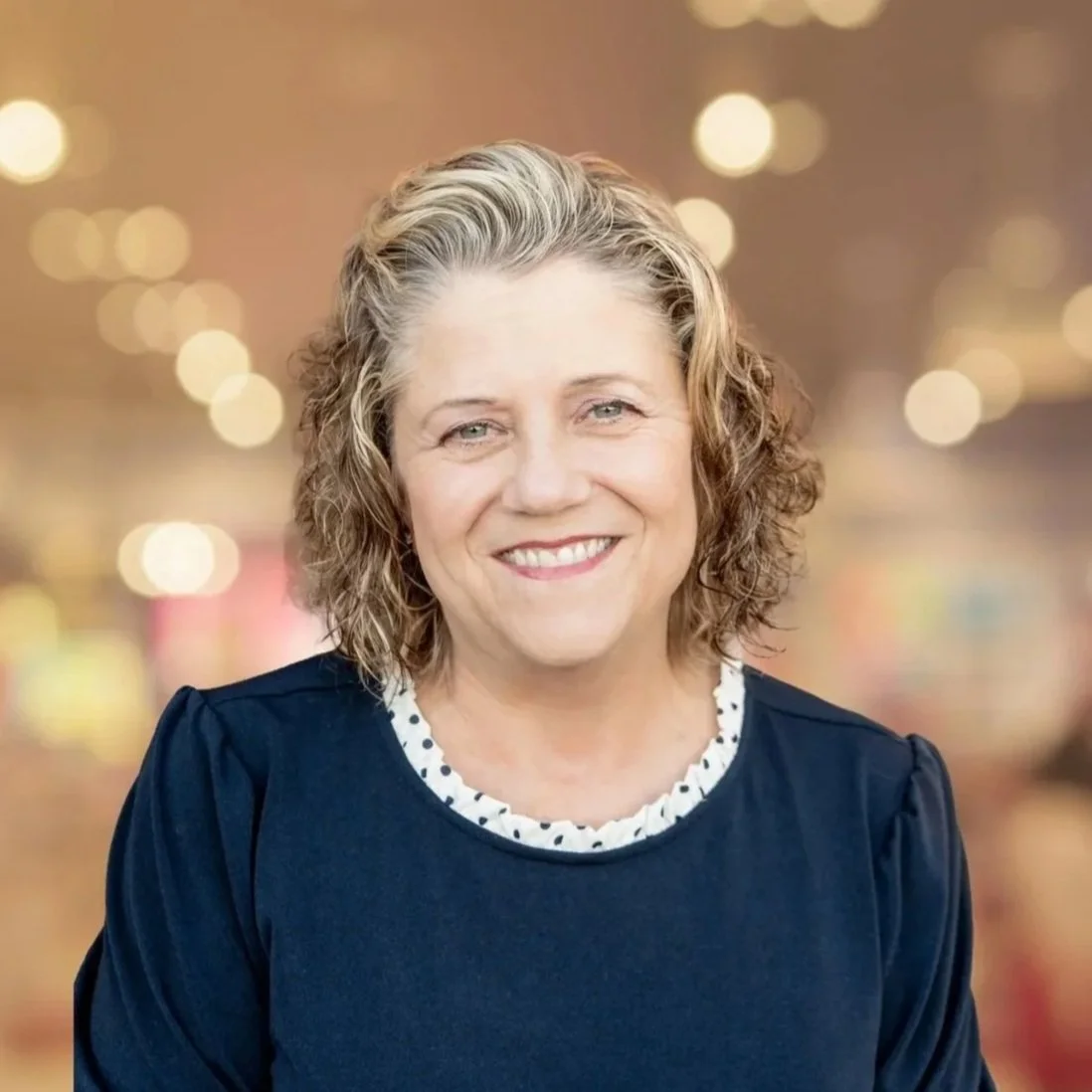 A smiling middle-aged woman with curly blonde hair sitting against a brick wall with fall-themed decorations in the background.