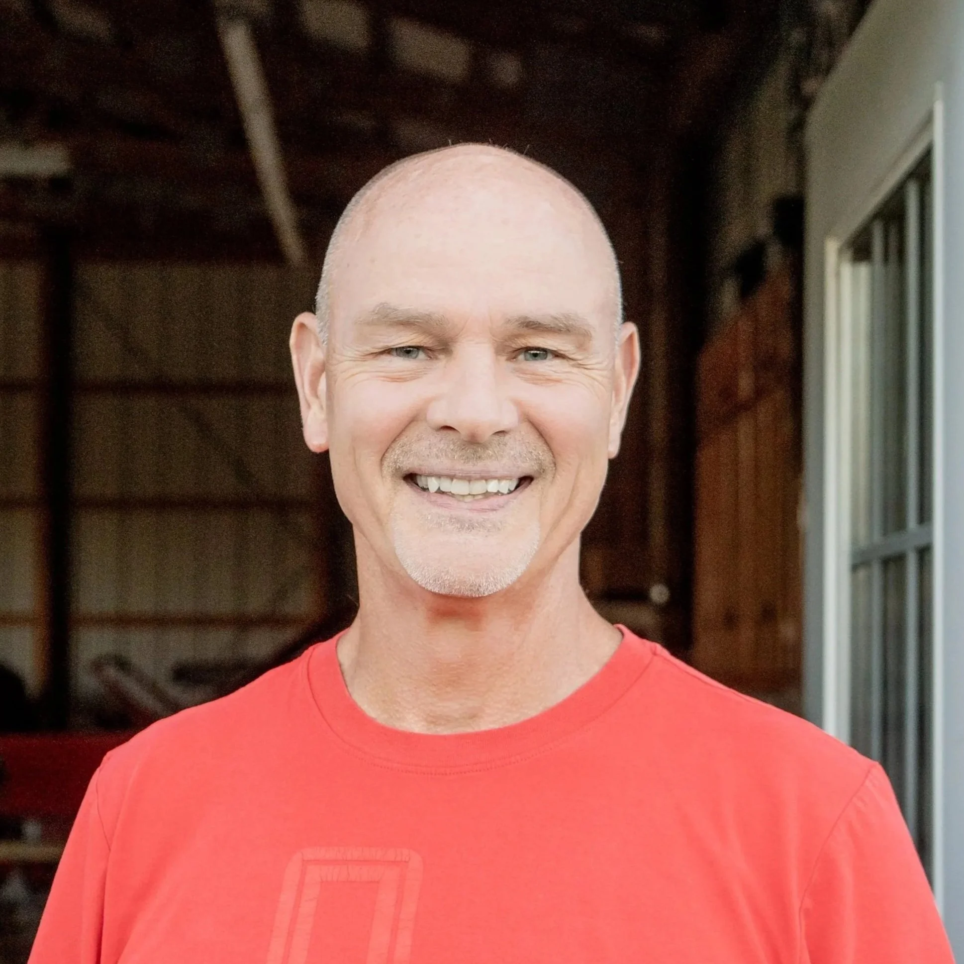 A smiling middle-aged man with a shaved head and light skin, wearing a red t-shirt, standing inside a wooden structure near a window.