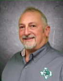 A smiling man with light brown hair dressed in a dark suit and bow tie, standing indoors against a neutral background.