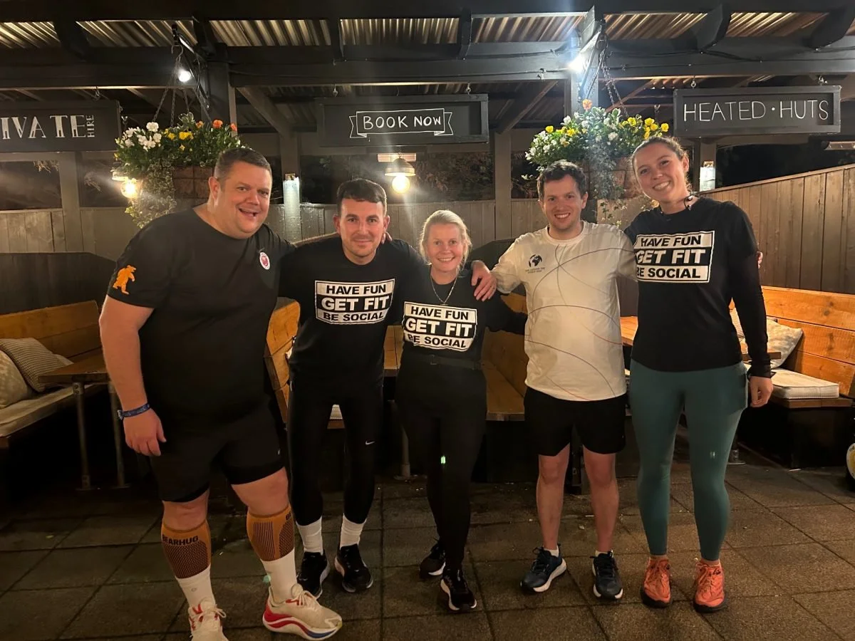 Group of five runners smiling and standing together in the beer garden of The Grove, Surbiton. Some wearing matching Pub run Club black T-shirts with the words "HAVE FUN, GET FIT, BE SOCIAL."