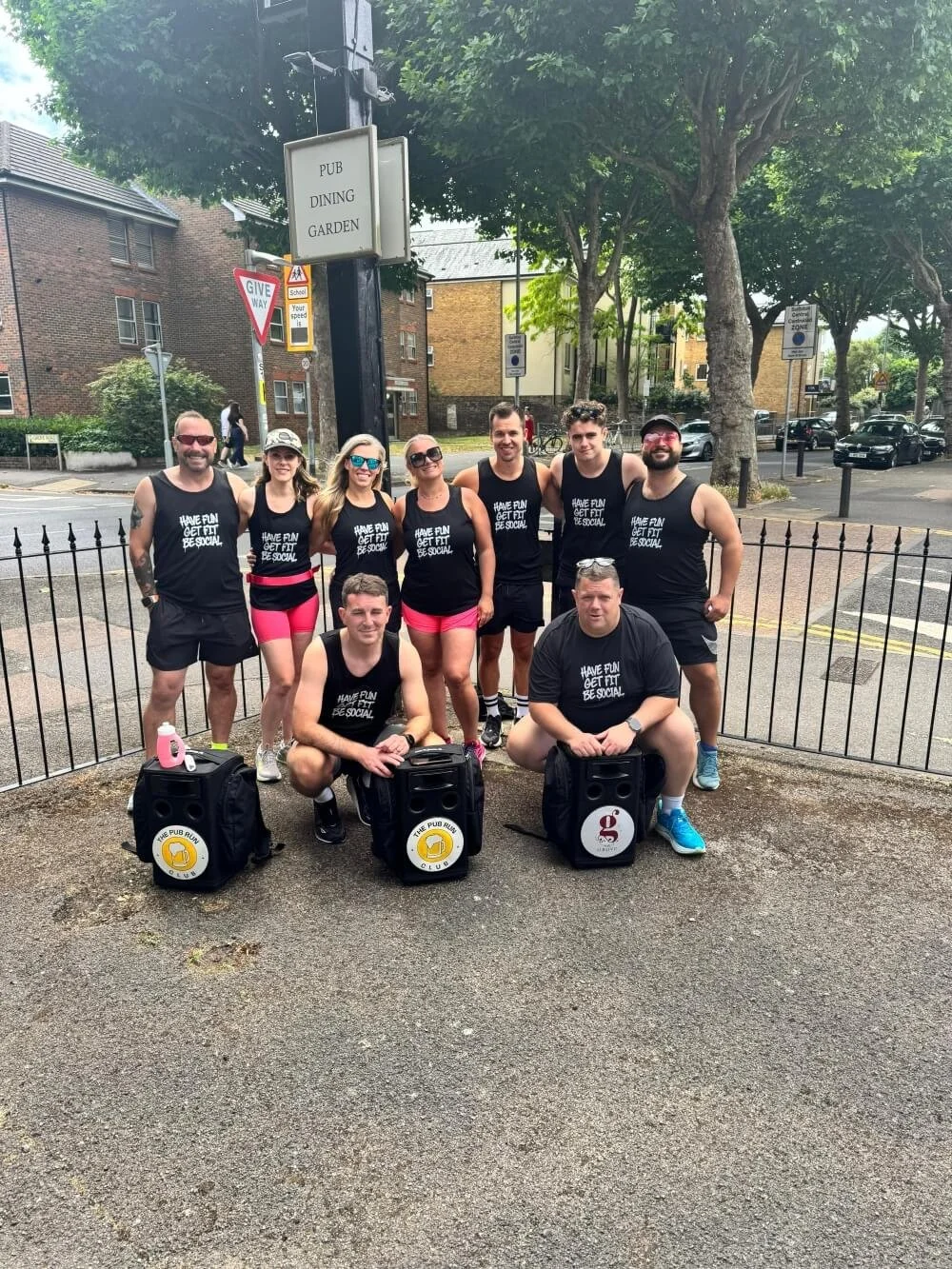 A group of nine runners wearing matching black tank tops with the text 'HAVE FUN, GET FIT, BE SOCIAL' on the front, posing outside The Grove Pub Surbiton. Some holding black speakers with yellow and red Pub Run Club labels.