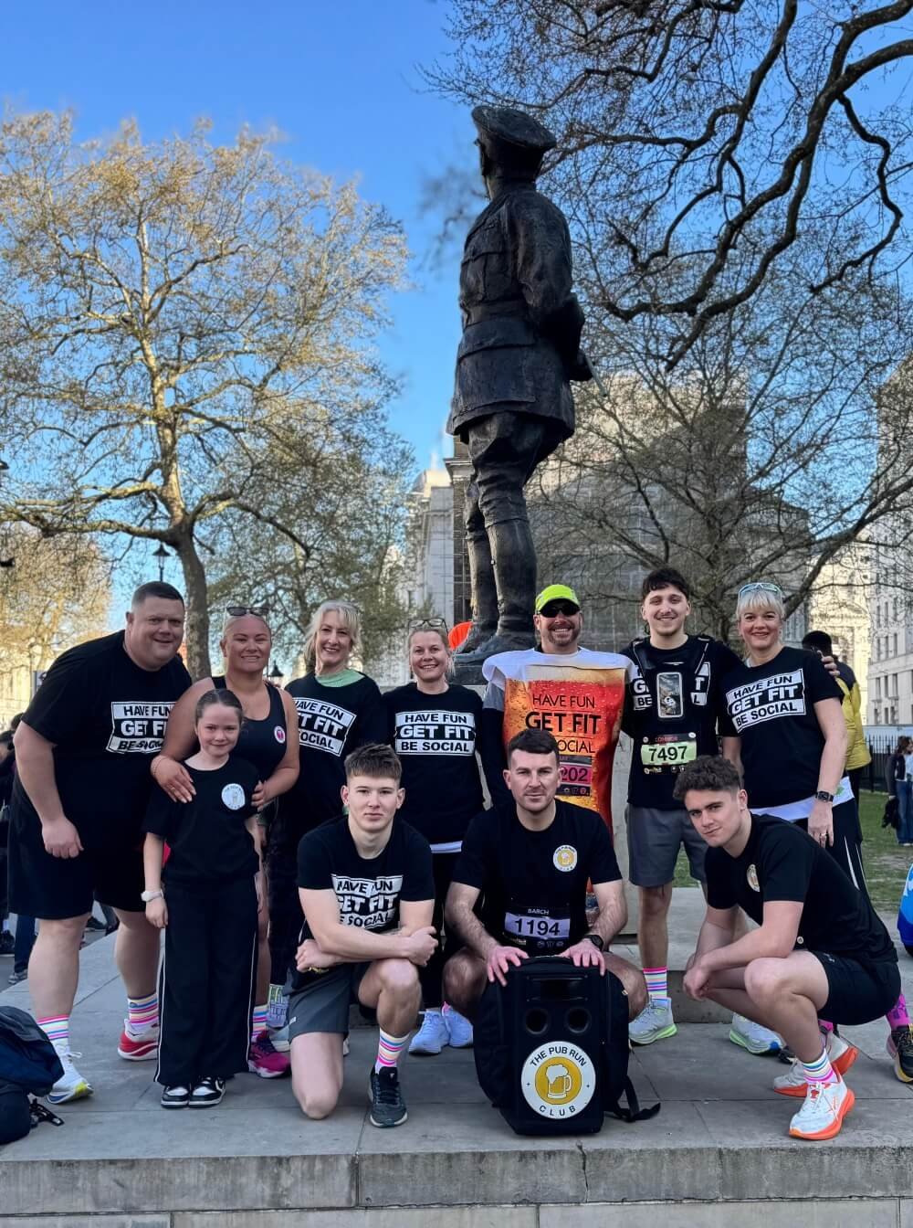Group of Pub Run Club runners wearing athletic clothing gathered in front of a statue in a park during a running event, with trees and buildings in the background.