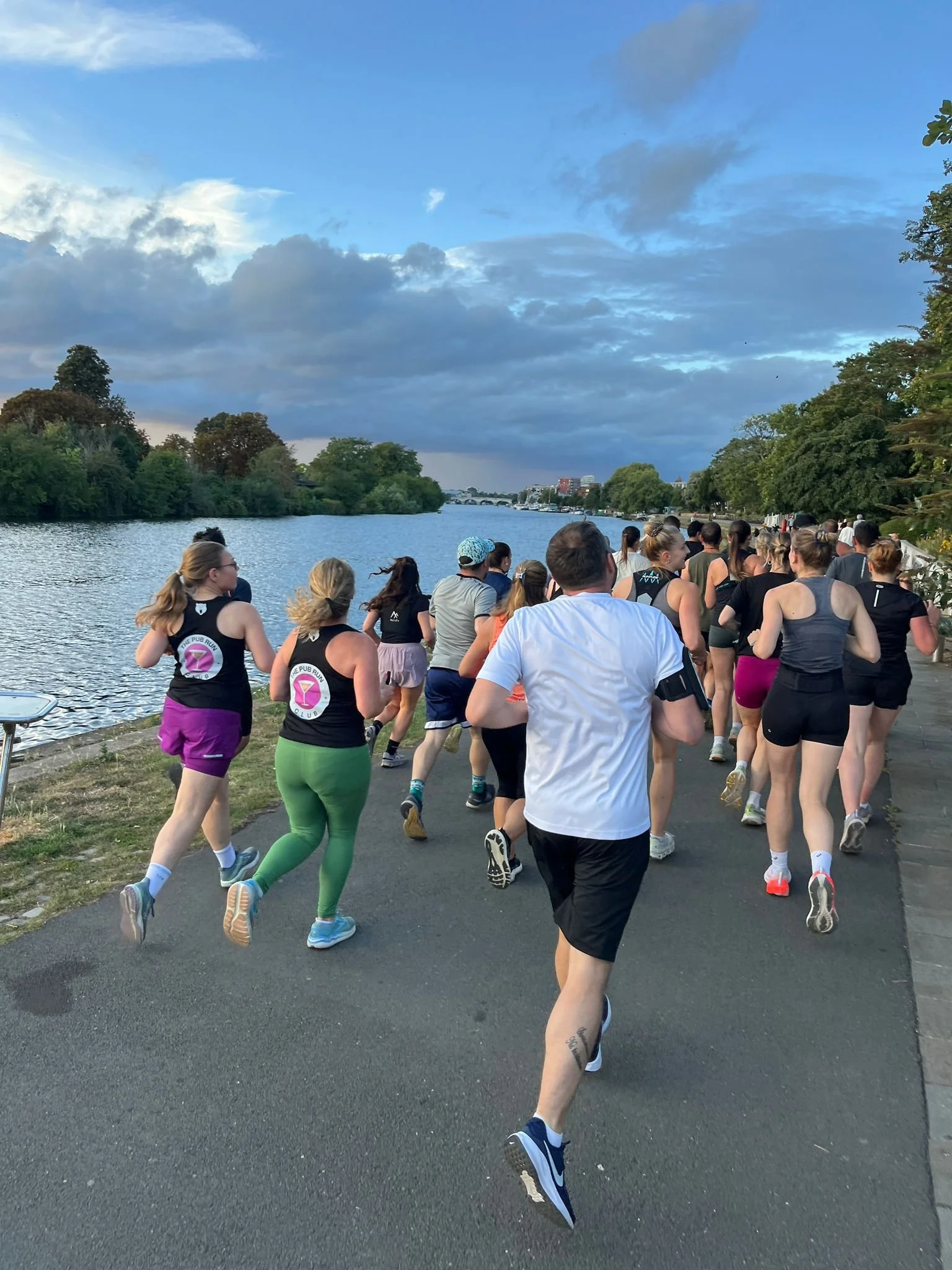 People running along a lakeside path during a race or marathon, with trees on both sides and a partly cloudy sky overhead.