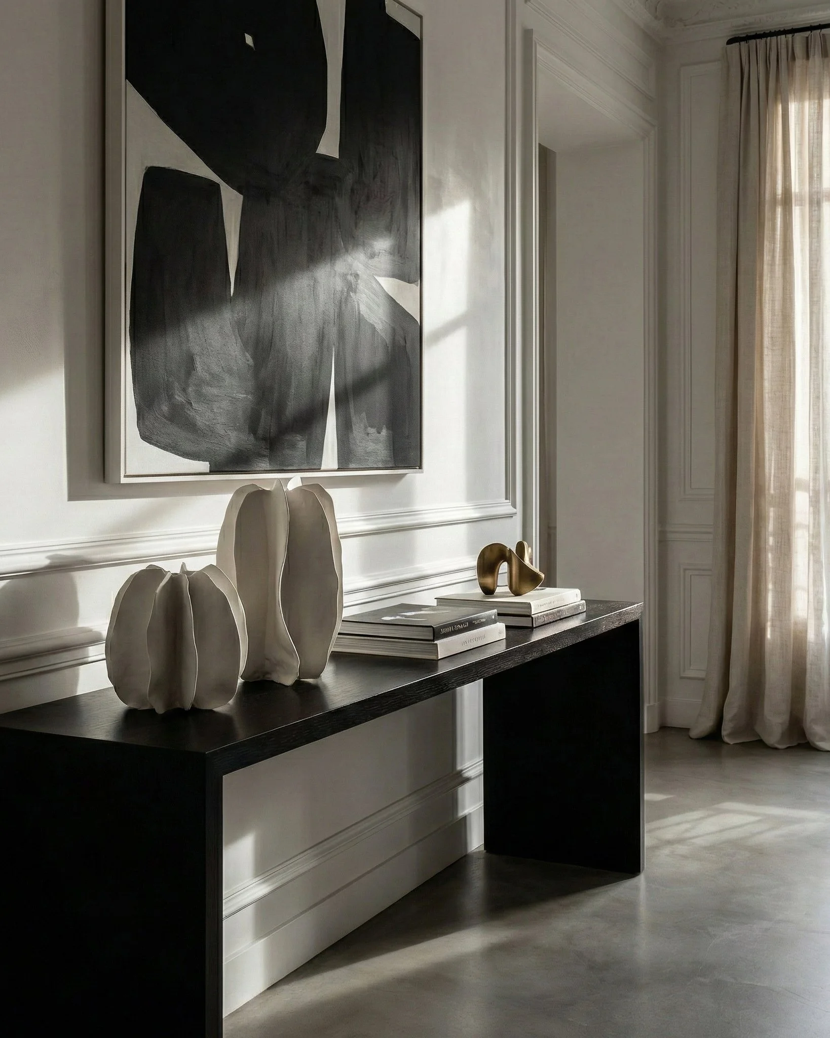 A black console table decorated with white ceramic vases, books, and a gold abstract sculpture, in front of a white wall with crown molding and a large black and white abstract painting.