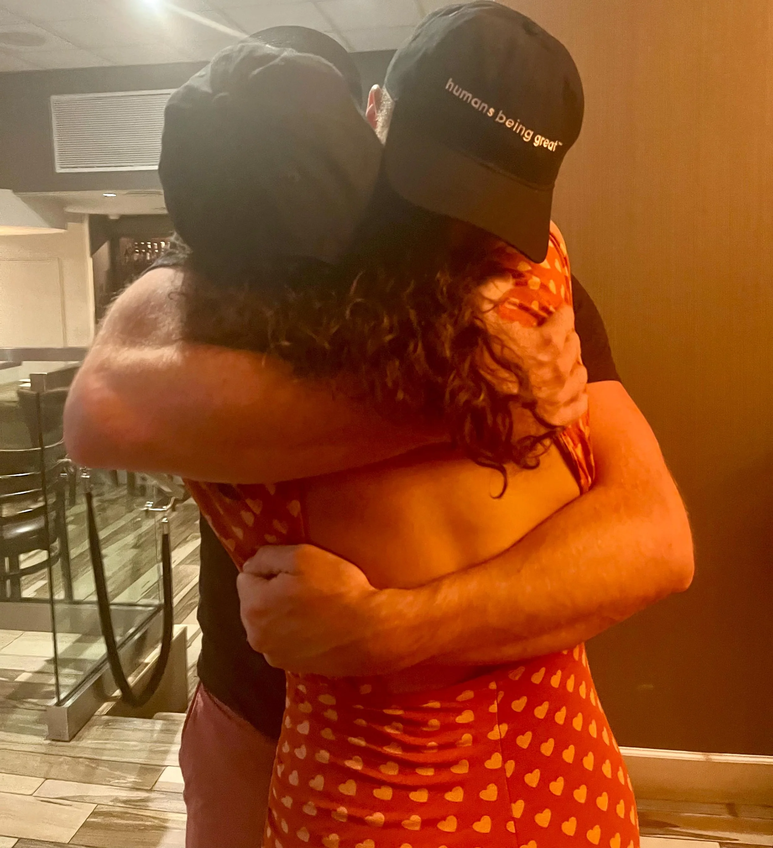 Two people embrace in a hug, one wearing a pink dress with orange hearts and a black cap with the slogan "humans being great," the other wearing a black shirt and shorts, in a room with wooden floors and a food service area in the background.